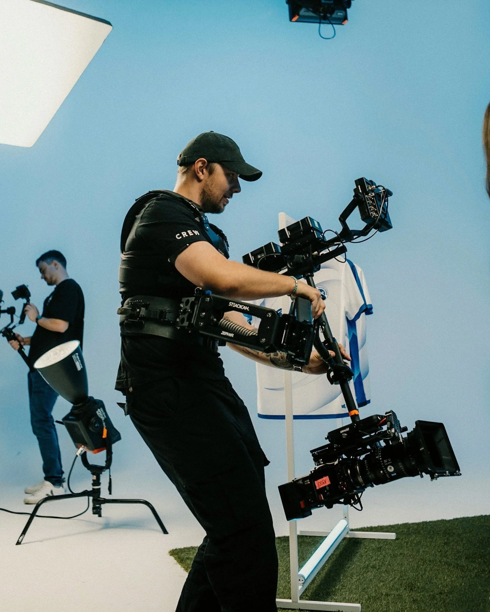 Filmteam bei der Arbeit im Studio, Person mit Kameraausrüstung - einer Steadicam, Personen mit technischer Ausrüstung, blauer Hintergrund, Studioausrüstung sichtbar.