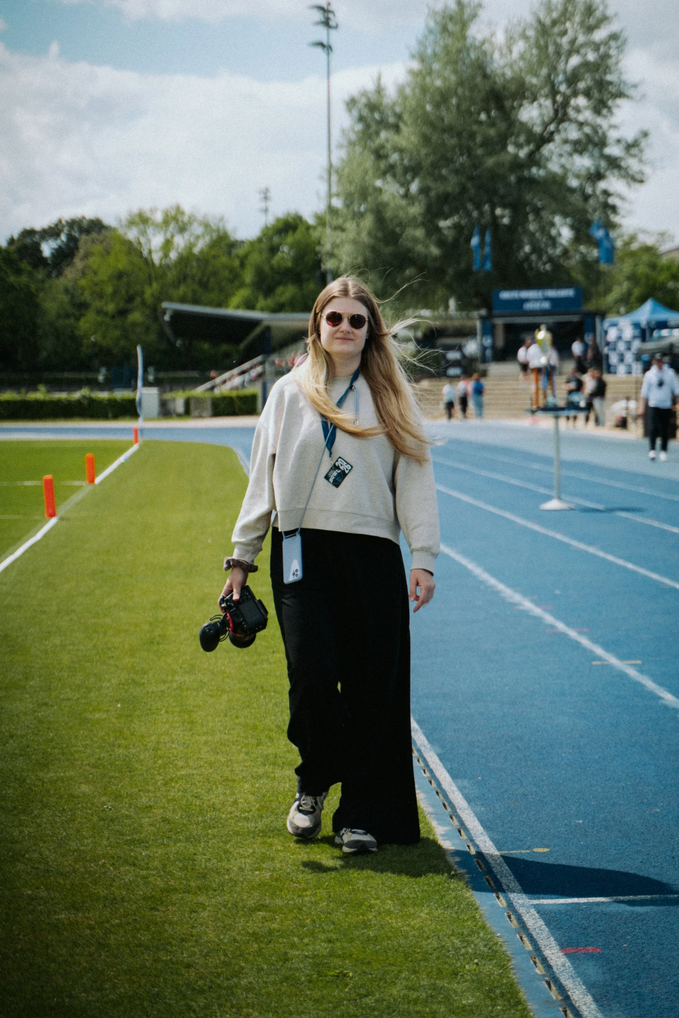 Eine junge Frau mit Sonnenbrille, langem blondem Haar und schwarzen Hosen, die auf einer Laufbahn im Stadion steht, umgeben von Menschen bei einer Veranstaltung - einem American Football Kids Camp.