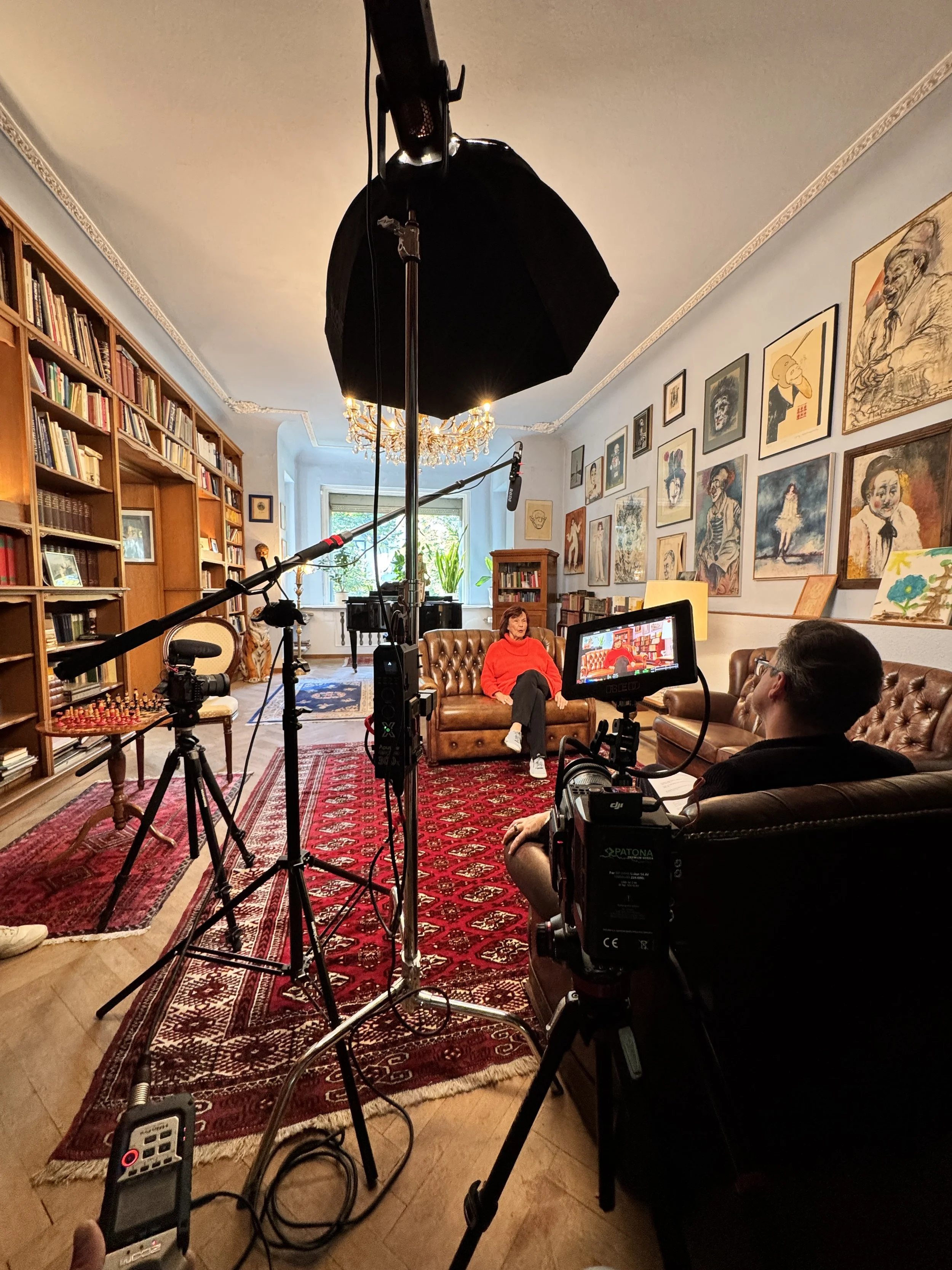 Ein Wohnzimmer mit einer Person in rotem Pullover, die auf einem braunen Ledersofa sitzt, umgeben von Kameras, Mikrofonen, einem Studio-Licht, Bücherregalen, Bildern an der Wand und einem Klavier im Hintergrund.
