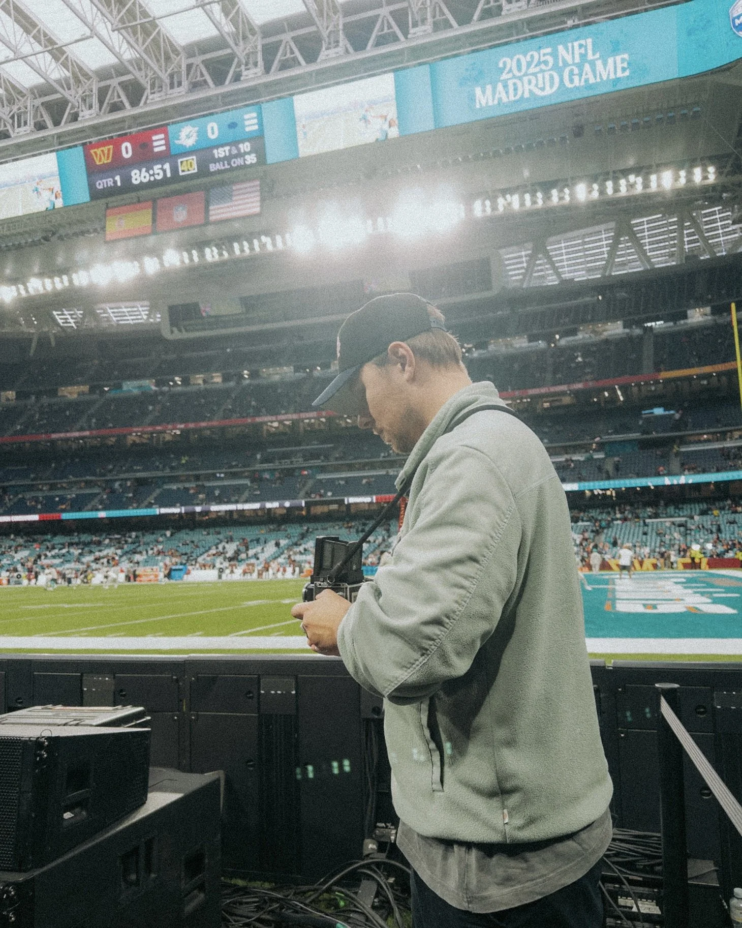 Ein Mann mit Kappe, der auf einem American Football Feld mit Fotografie-Equipment - einer Rolleiflex - steht, im Hintergrund eine große LED-Leinwand im Stadion mit Annotations zur NFL-Spiele 2025 in Madrid.