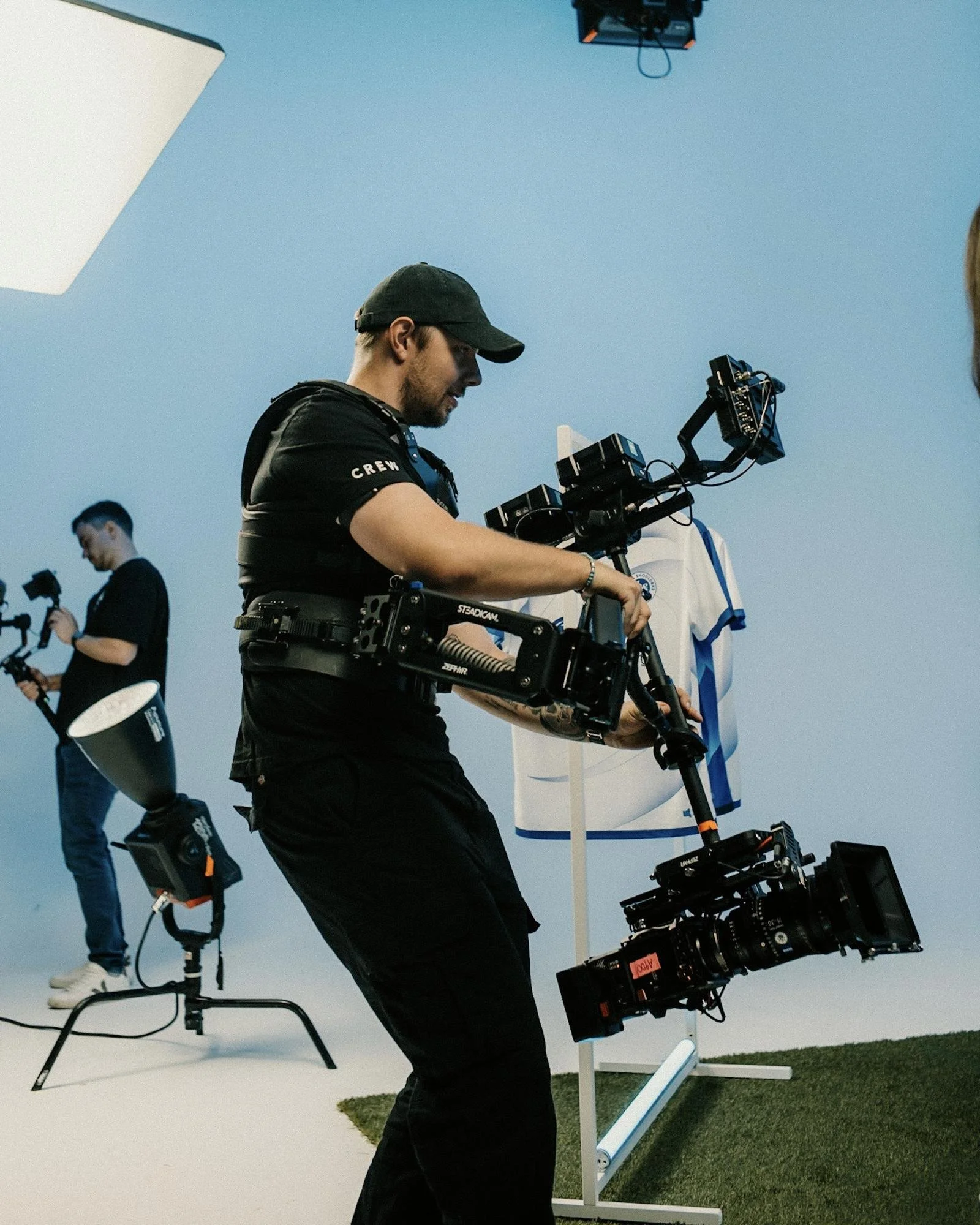 Filmteam bei der Arbeit im Studio, Person mit Kameraausrüstung - einer Steadicam, Personen mit technischer Ausrüstung, blauer Hintergrund, Studioausrüstung sichtbar.
