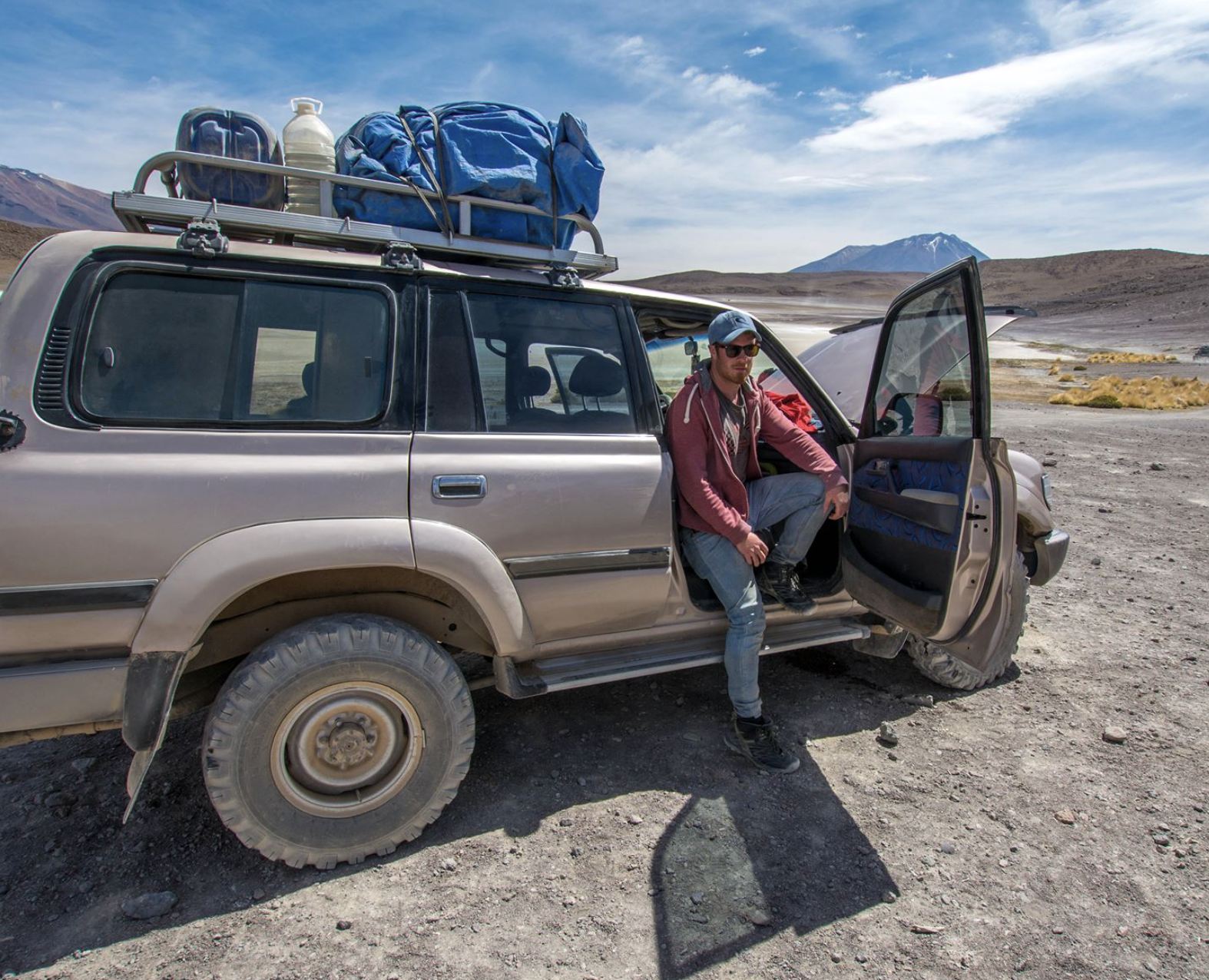 A man sitting in the open door of a muddy beige SUV with a loaded roof rack, in a barren desert landscape with mountains and a partly cloudy sky in the background.