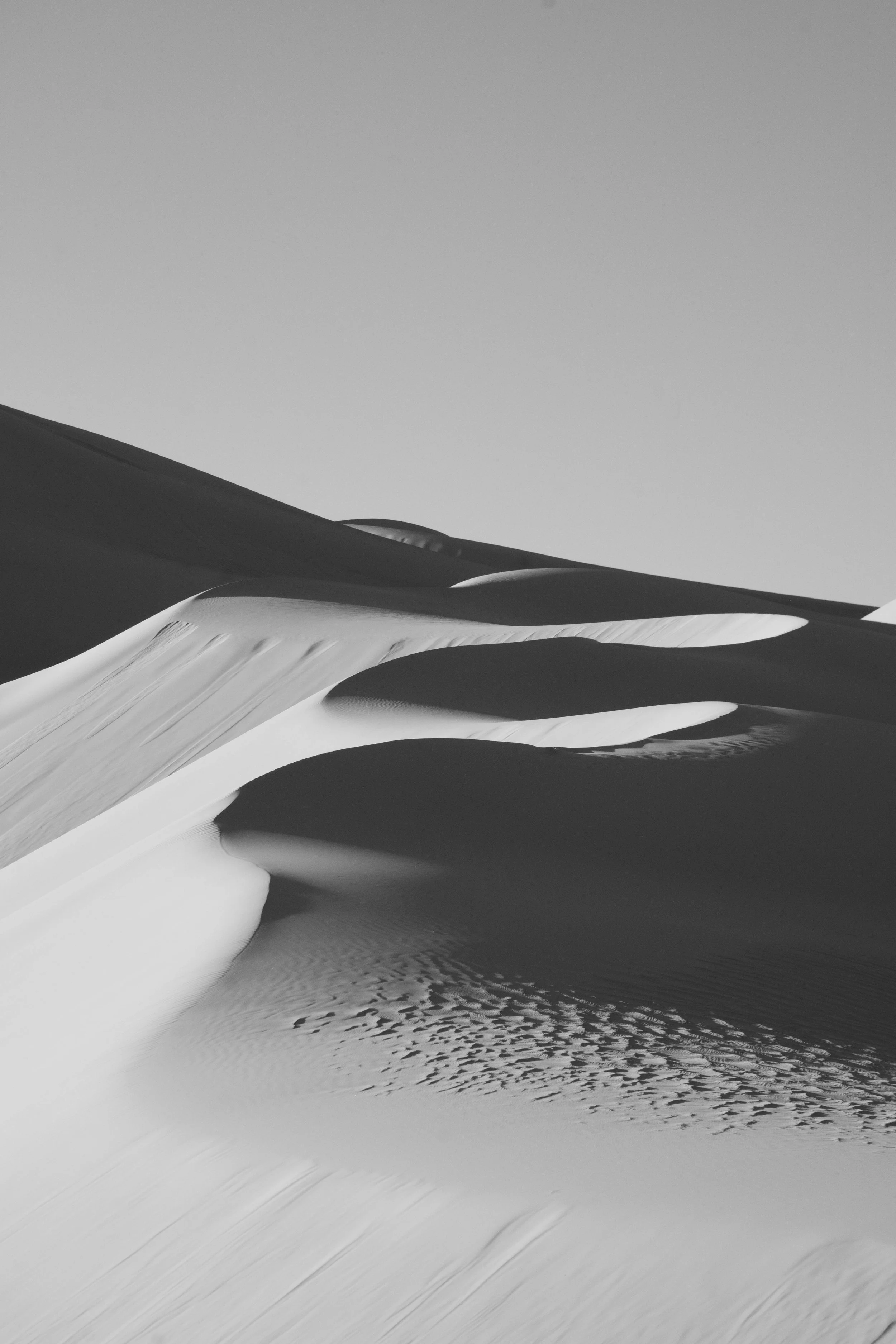 Black and white photo of sand dunes with smooth curves and ripples, under a clear sky.