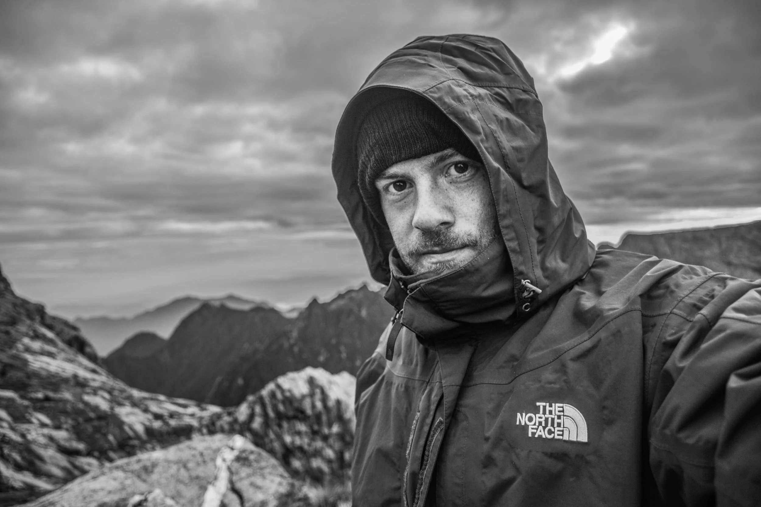 A man wearing a jacket and beanie outdoors, taking a selfie with mountain scenery in the background, in black and white.