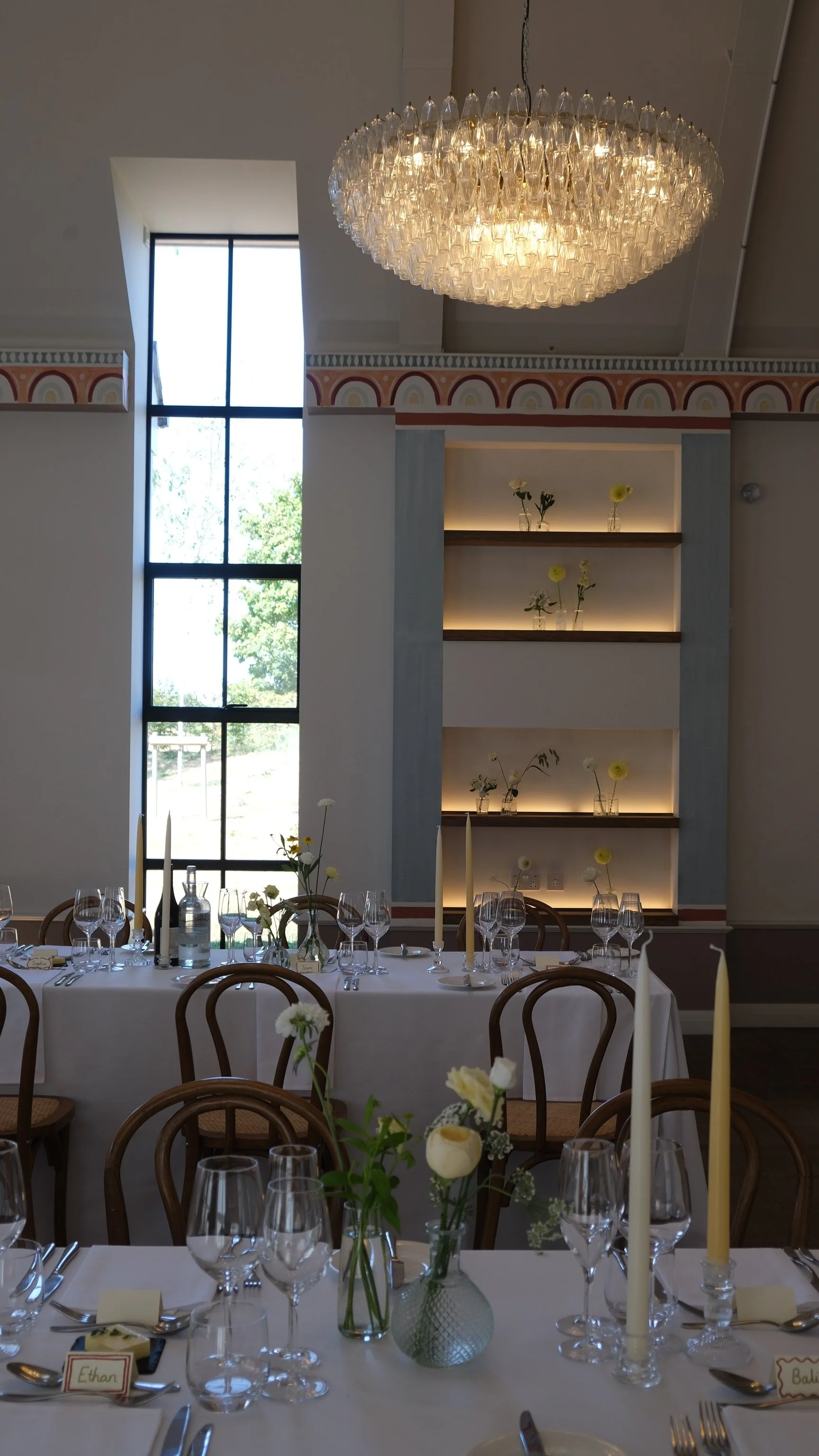 Elegant dining table set with glassware, candles, and floral centerpiece in a room with large window and chandelier.