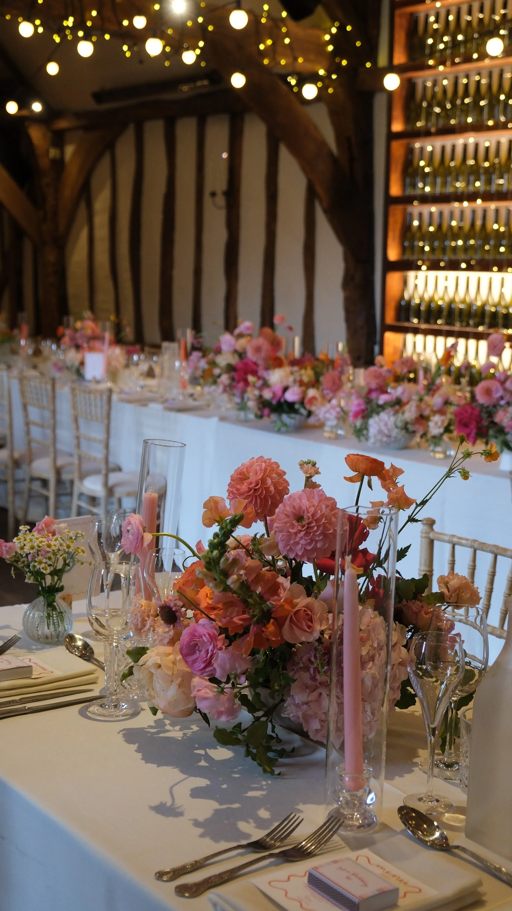Elegant wedding reception table decorated with pink and peach flowers, white candles, and fine glassware, set in a rustic barn with wood beams and string lights.