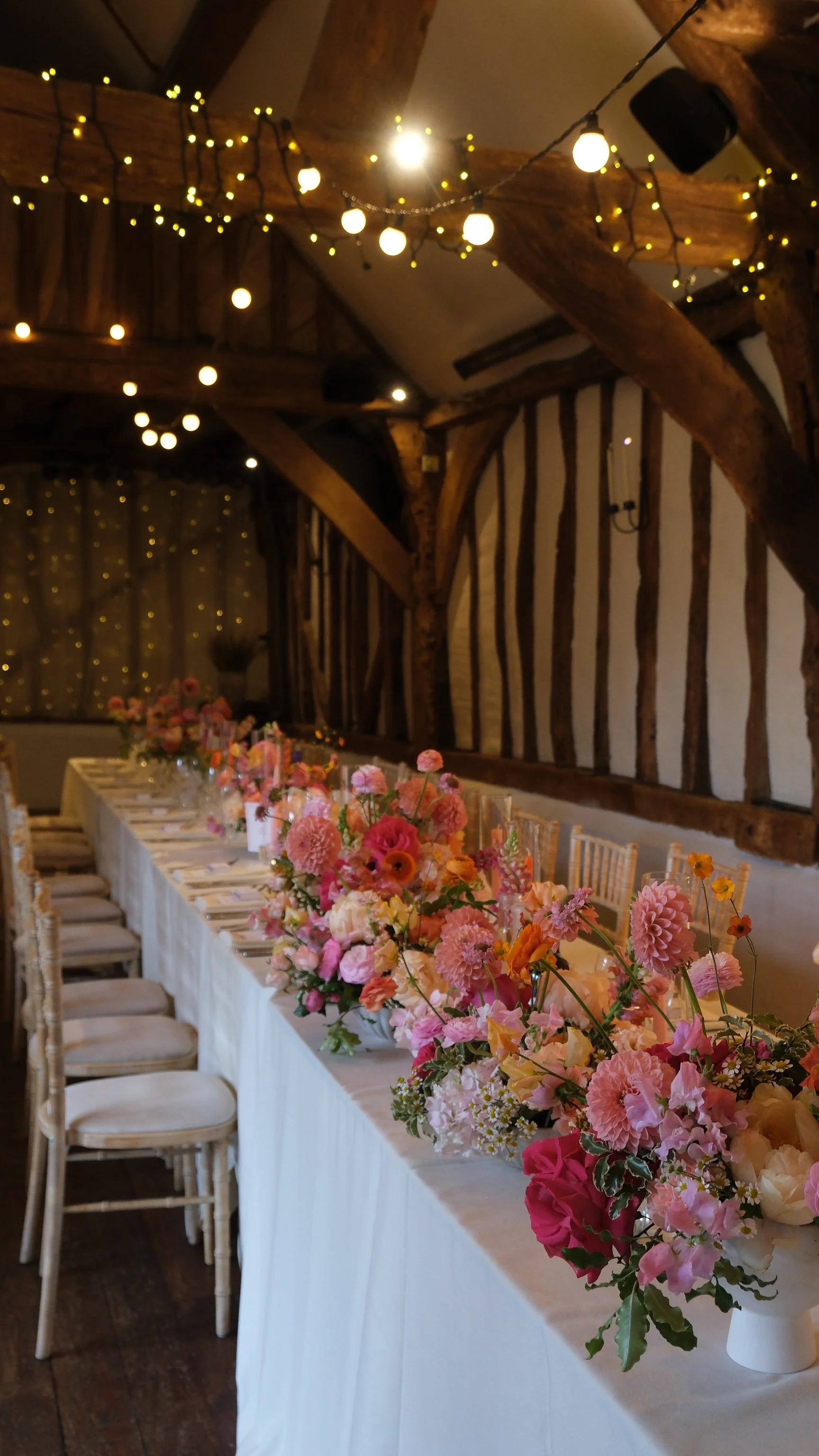A long banquet table decorated with pink and peach flowers and place settings, inside a rustic venue adorned with string lights and wooden beams.