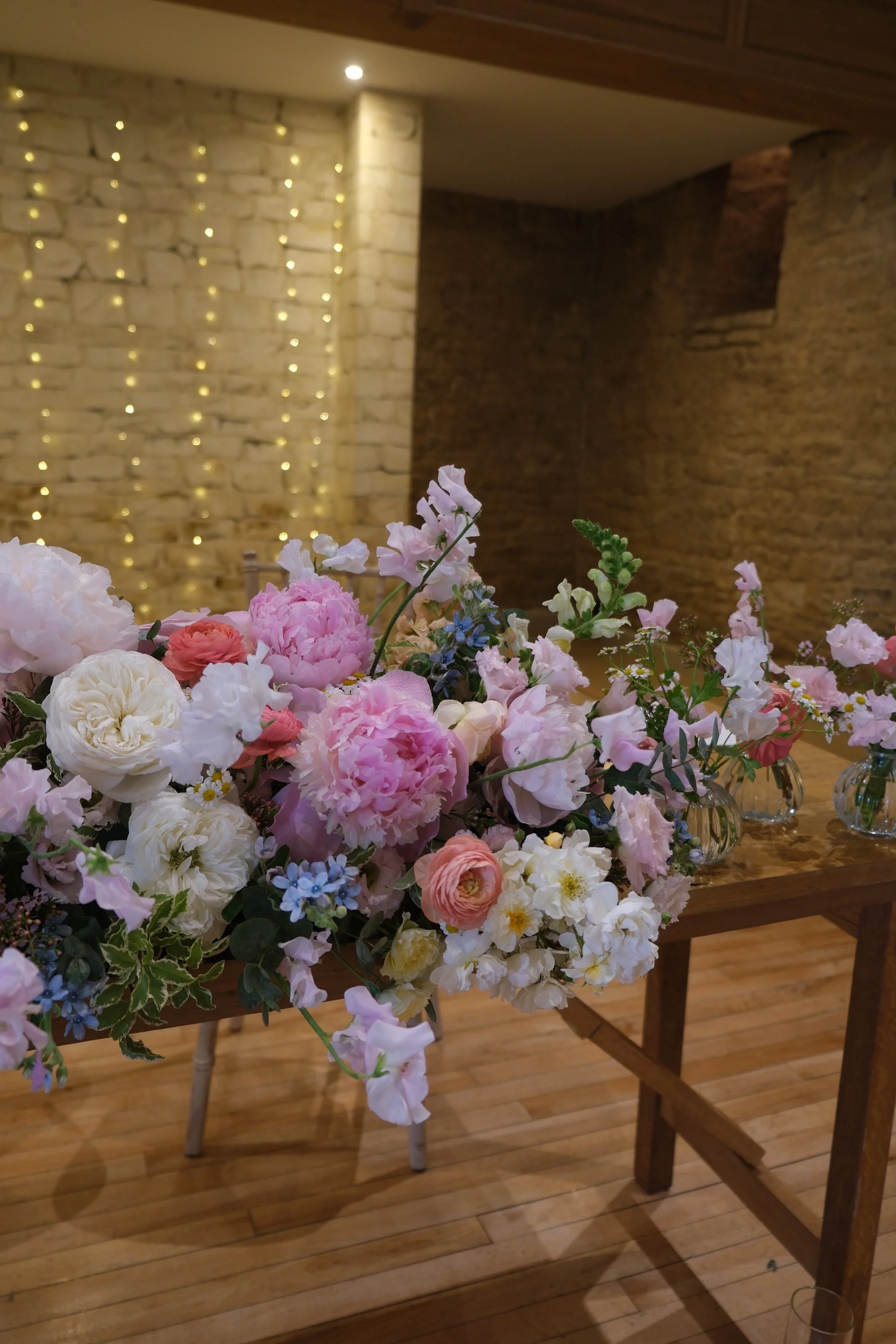 A colorful bouquet of various flowers including pink, white, and light purple blooms, arranged on a wooden table indoors. In the background, there is a brick wall with string lights hanging and wooden cabinetry on the ceiling.