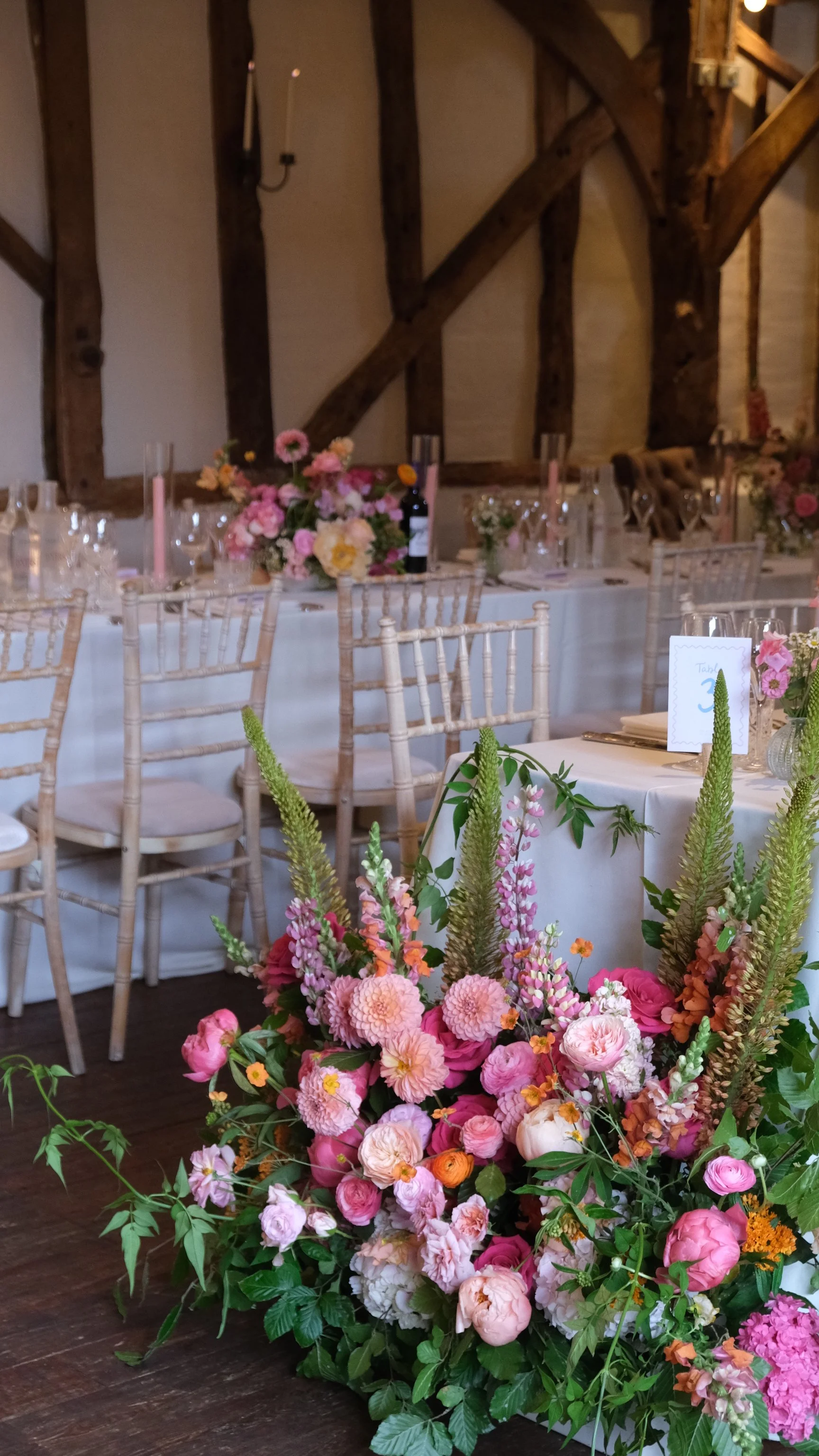 A wedding or event reception decorated with pink and peach flowers, tall floral arrangements, and table settings, with wooden chairs and rustic beams on the ceiling.