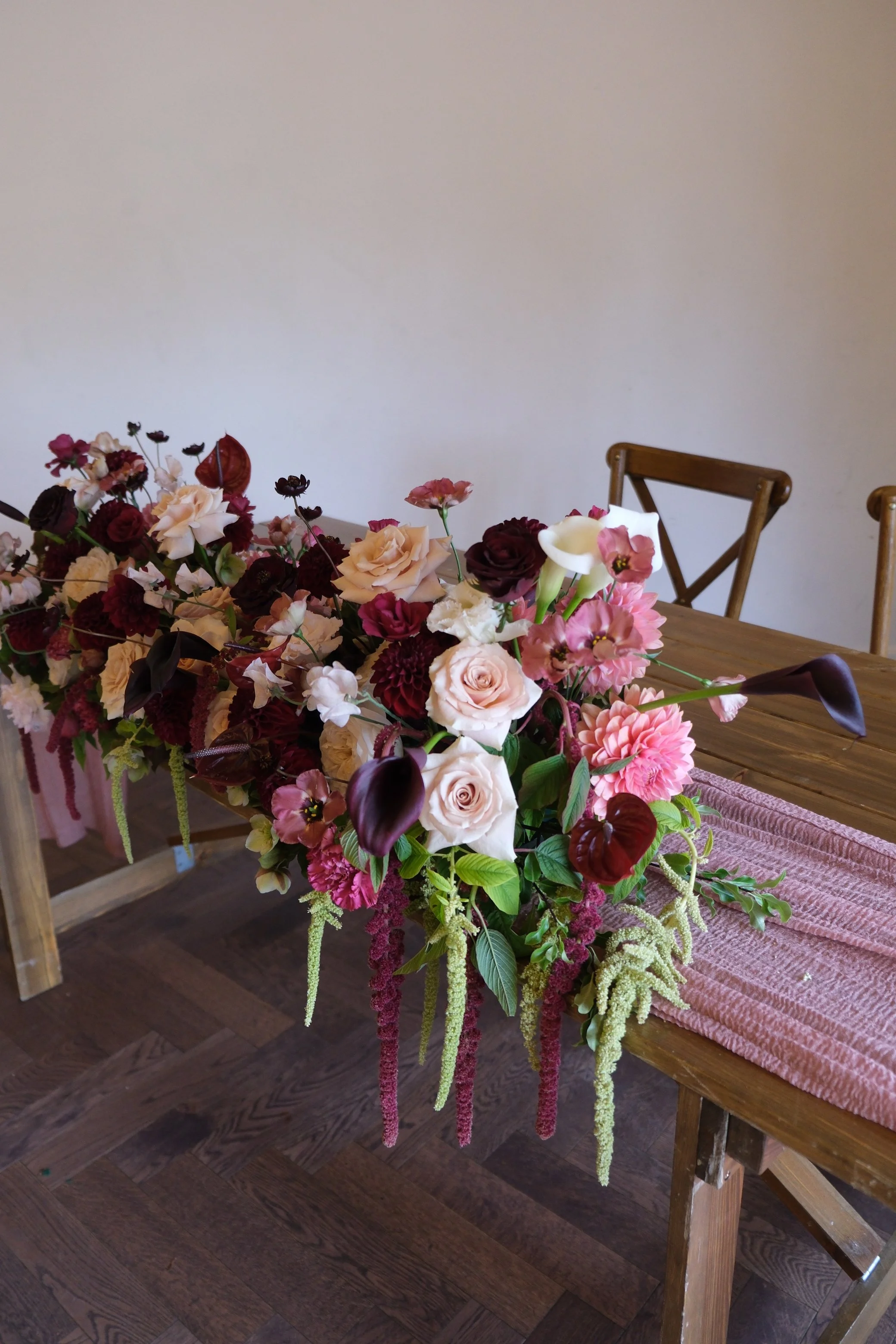 A wooden table decorated with an elaborate floral arrangement featuring various pink, white, and dark red flowers. The table has a pink textured cloth and is positioned in a room with wooden flooring and wooden chairs in the background.
