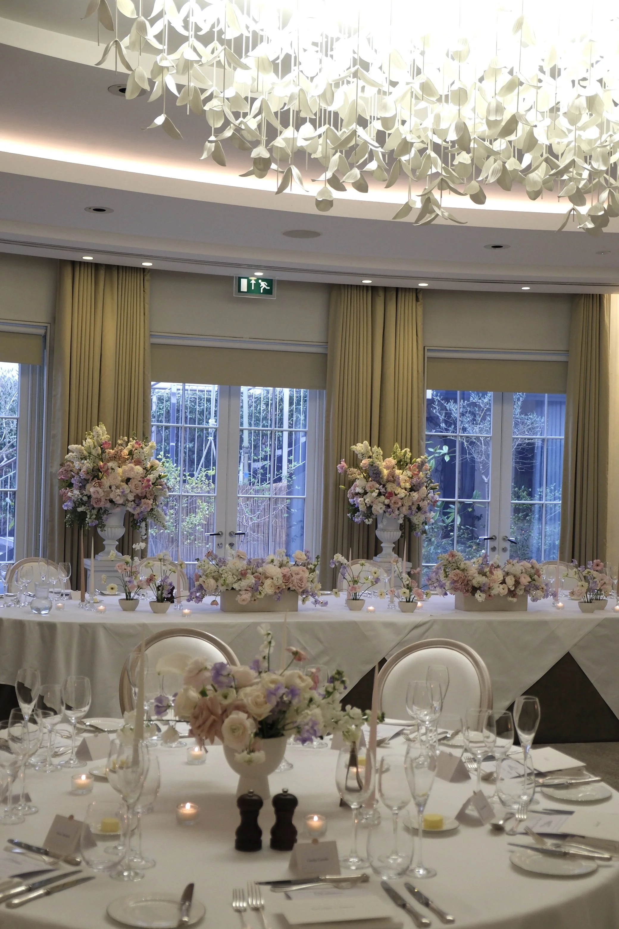 Elegant indoor dining area decorated for a wedding or special event, with round tables covered in white tablecloths, floral centerpieces, and lit candles. Floor-to-ceiling windows with beige curtains reveal a garden outside. A large floral chandelier hangs above.