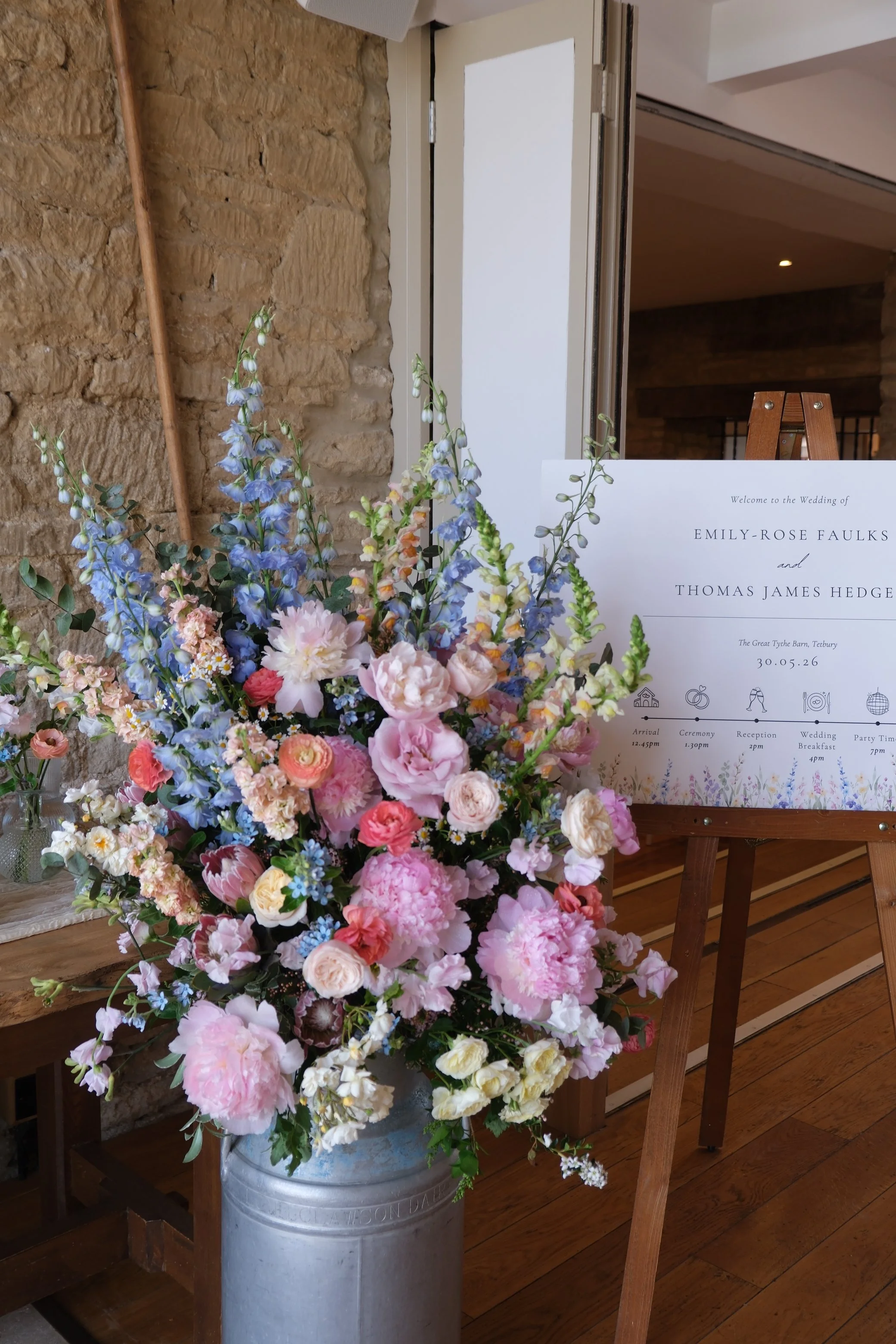 A large bouquet of pink, white, and purple flowers in front of a wedding sign with details about the wedding of Emily-Rose Faulks and Thomas James Hedge at The Great Tythe Barn, Tetbury, on May 30, 2026. The sign includes a schedule and icons for arrival, ceremony, reception, wedding breakfast, and party.
