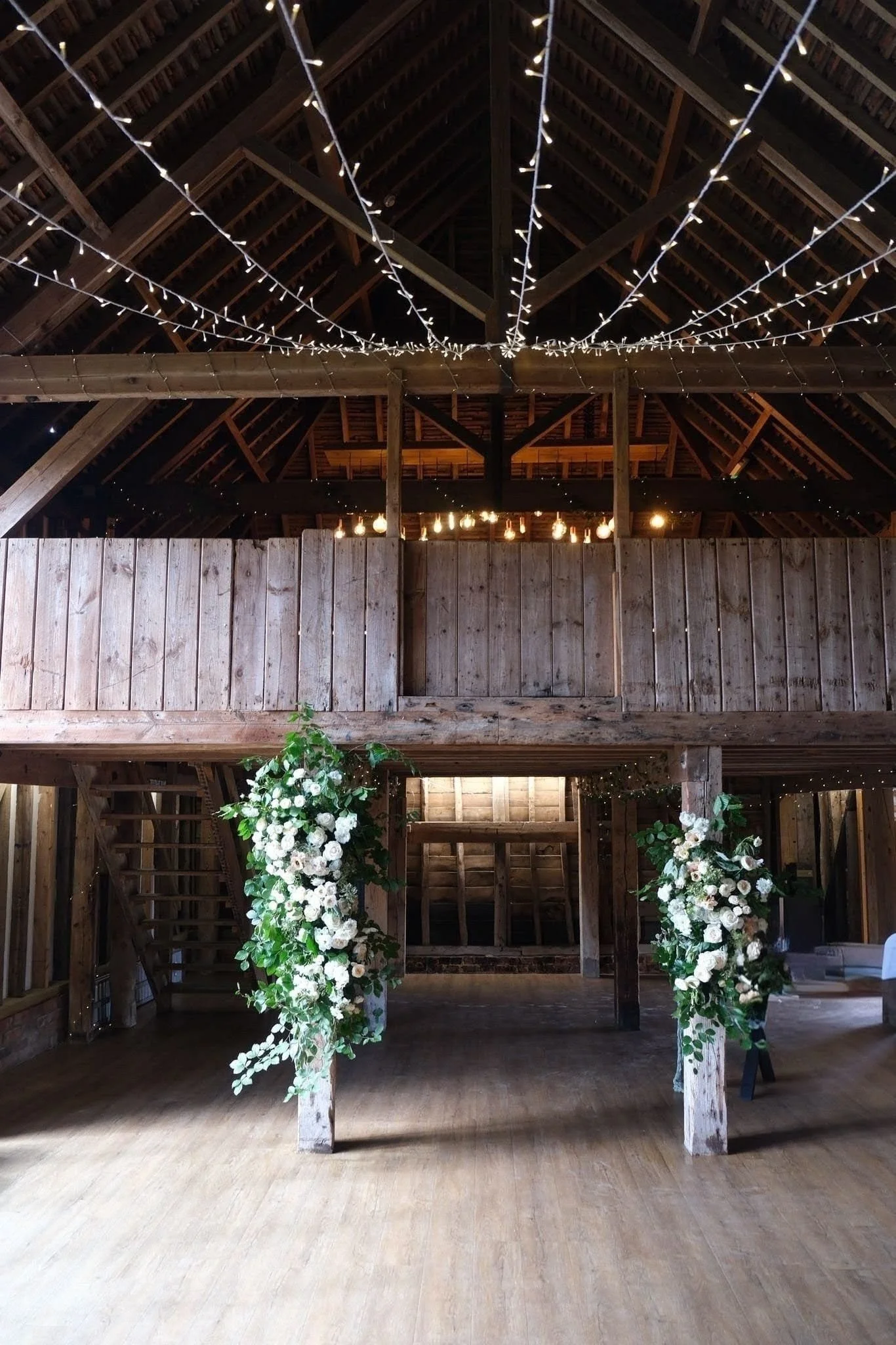 Interior of a rustic barn with wooden beams and string lights, decorated with white floral arrangements at the entrance, likely for a wedding or event.