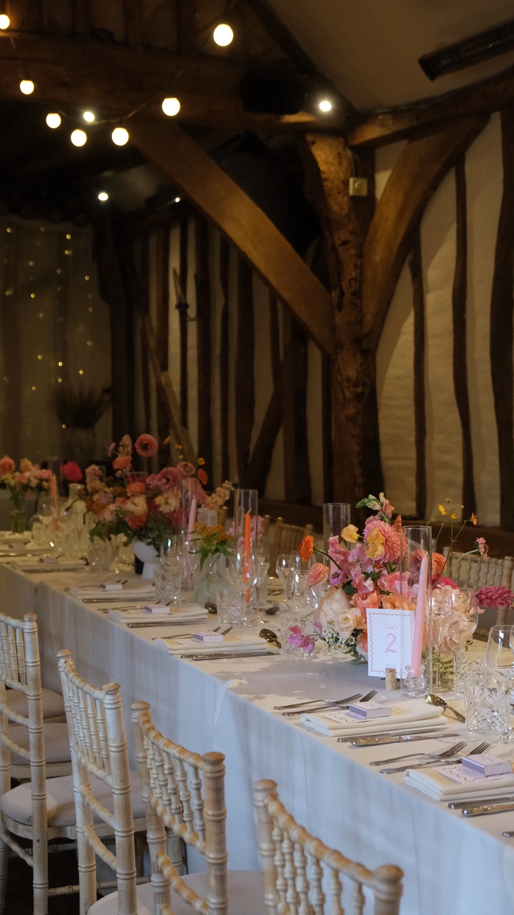 A long banquet table decorated with pink and peach flowers, set with beads, glassware, and napkins in a rustic room with wooden beams and string lights.