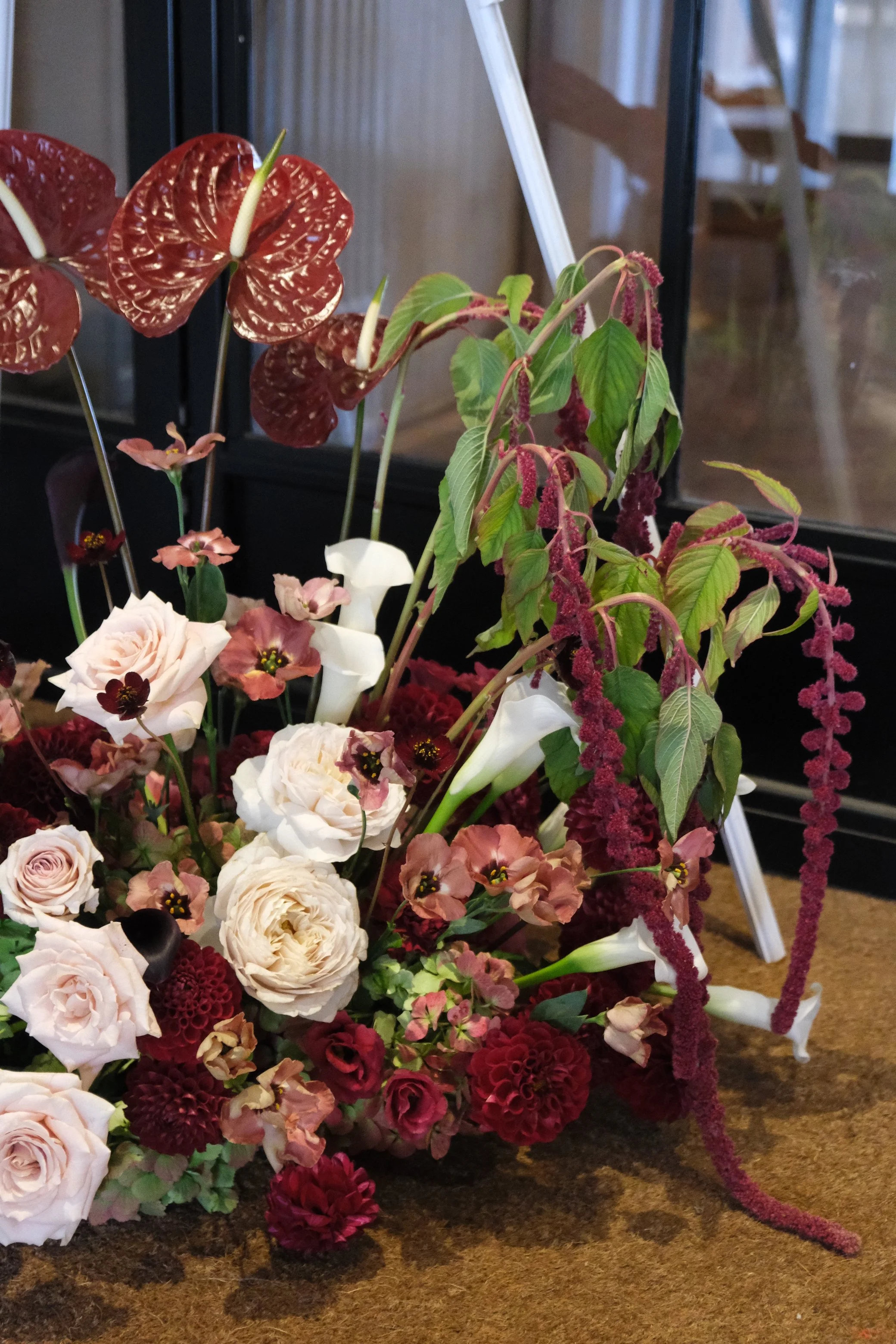 A floral arrangement with pink and red flowers, green leaves, and some plants with red and white spathiphyllum leaves, placed on a brown surface against a background of black and glass windows.