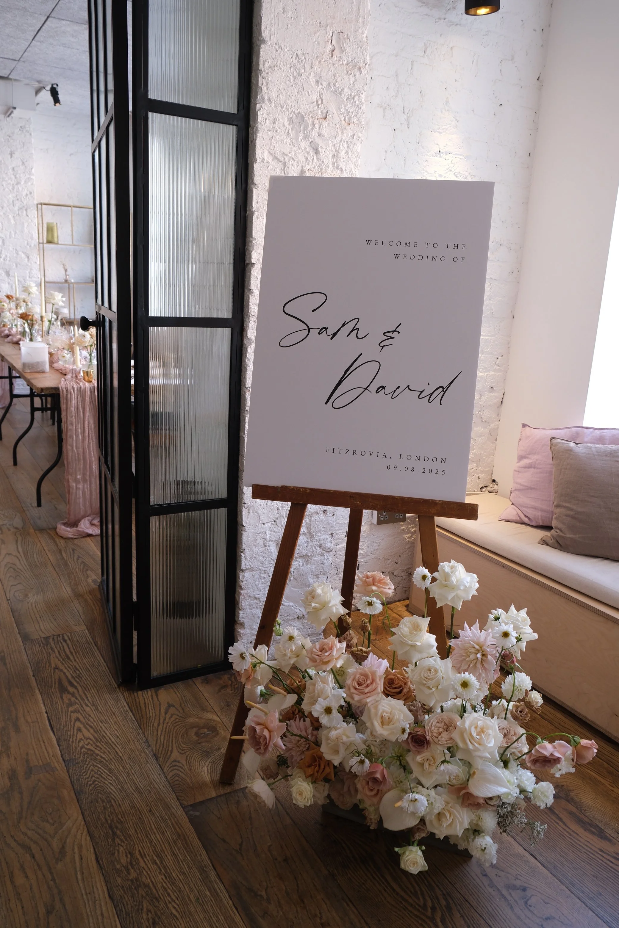 A wedding welcome sign with the names 'Sam & David' and the date '09.08.2025', surrounded by a flower arrangement with white, blush, and peach roses, placed on a wooden easel inside a decorated venue with white brick walls, a pink cushioned bench, and floral decor.