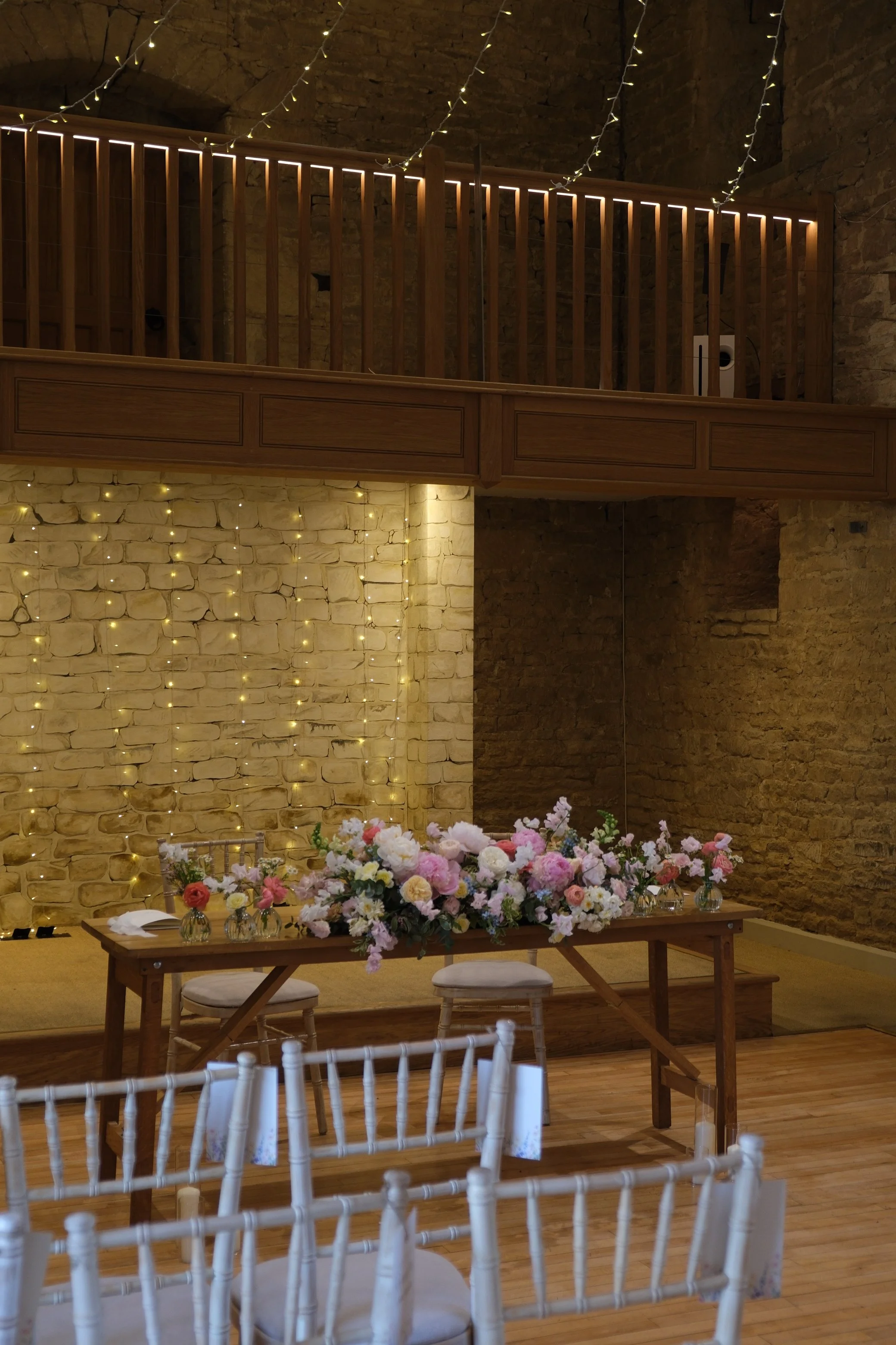 Indoor wedding reception setup with white chairs, a long wooden table with a large floral centerpiece, fairy lights on the brick wall, and a wooden balcony with string lights.