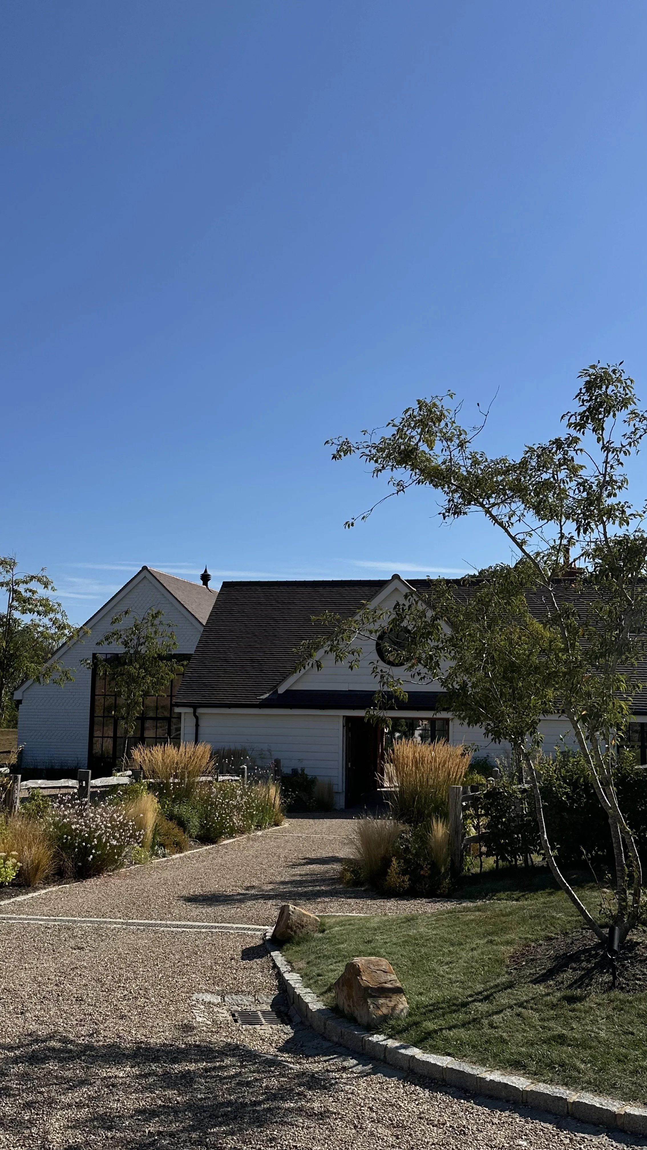 A house with a white exterior and a dark brown roof, surrounded by a garden with trees, bushes, and ornamental grasses. The sky is clear and blue.