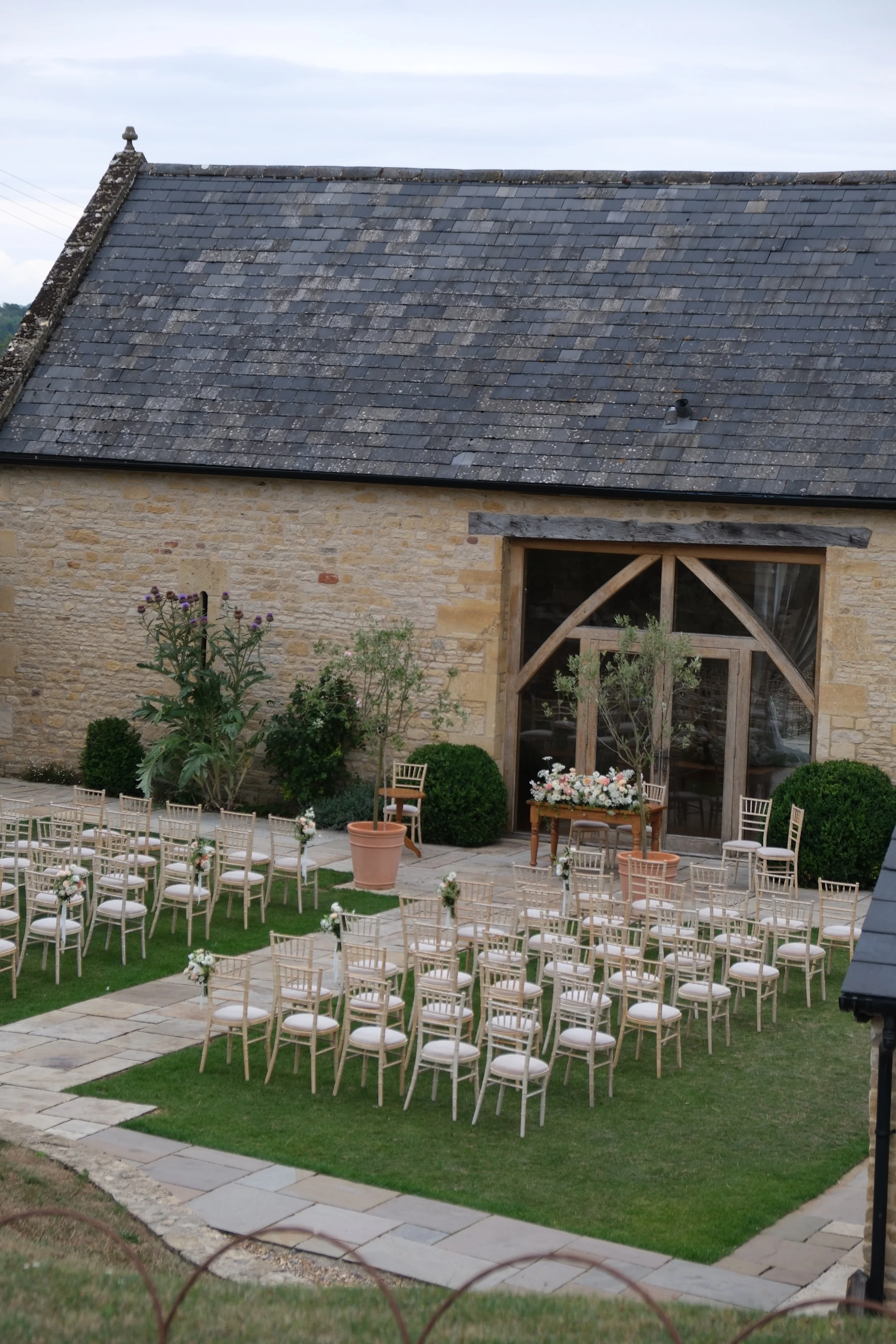 An outdoor wedding setup in front of a rustic stone building with covered wooden beams. There are rows of white chairs arranged on a grassy lawn with white floral decorations, and a small table with flowers near the front. The background shows a stone wall with a large window.