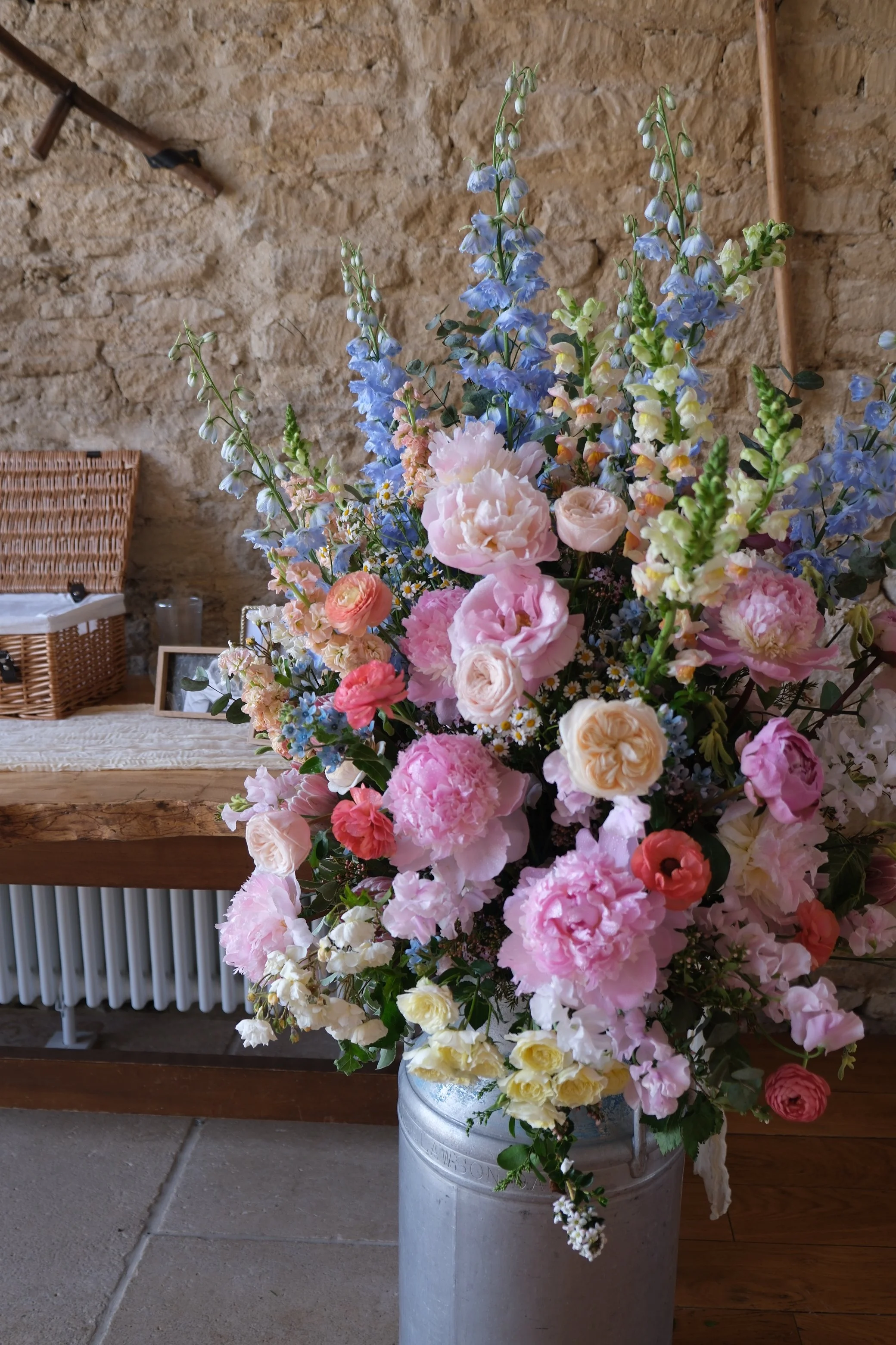 A colorful bouquet of flowers in a silver metal bucket in front of a rustic stone wall and a wooden table with wicker basket, framed photo, and glass jars.