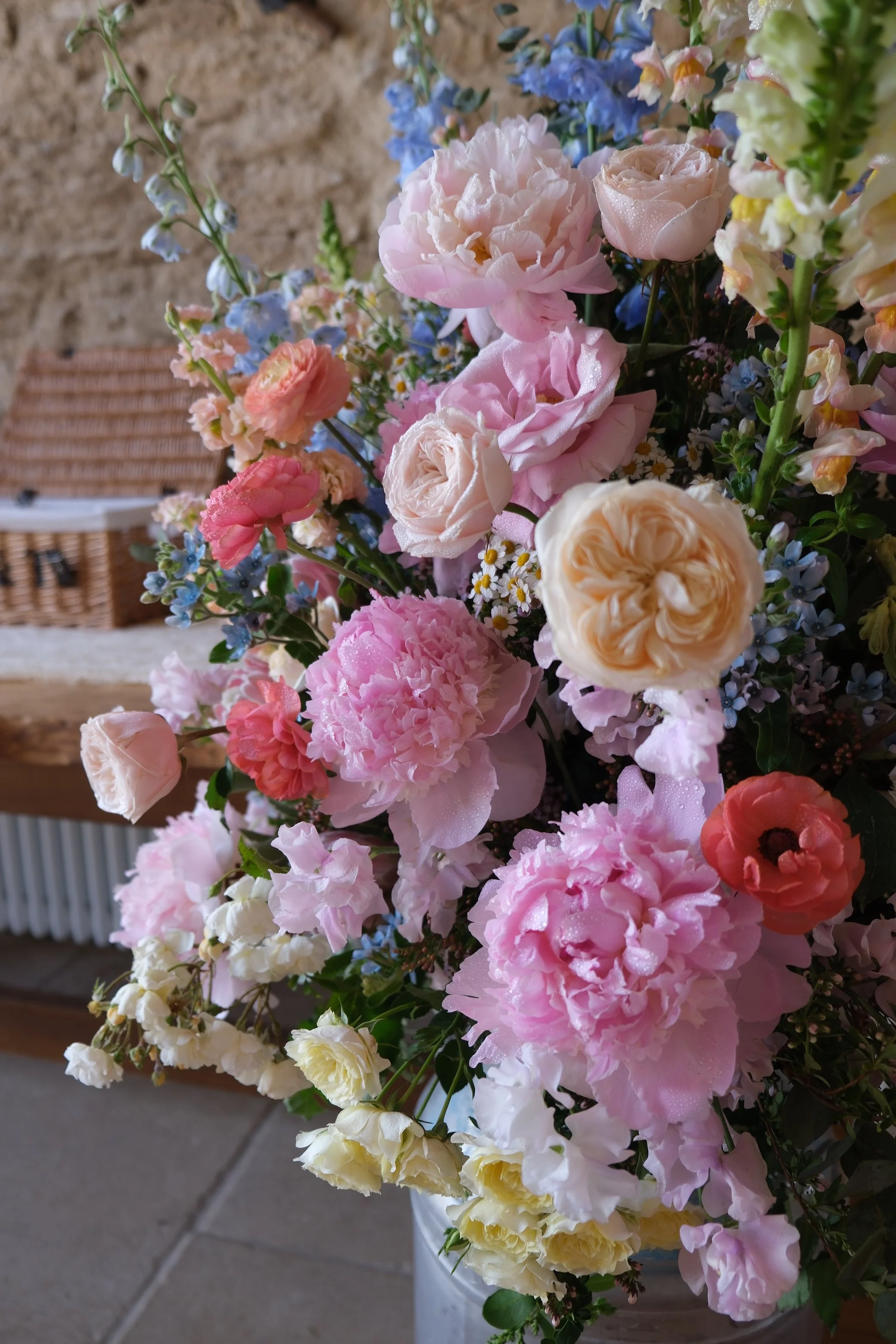 A colorful arrangement of flowers, including pink, cream, yellow, and blue blossoms, set against a rustic wooden background with a basket nearby.