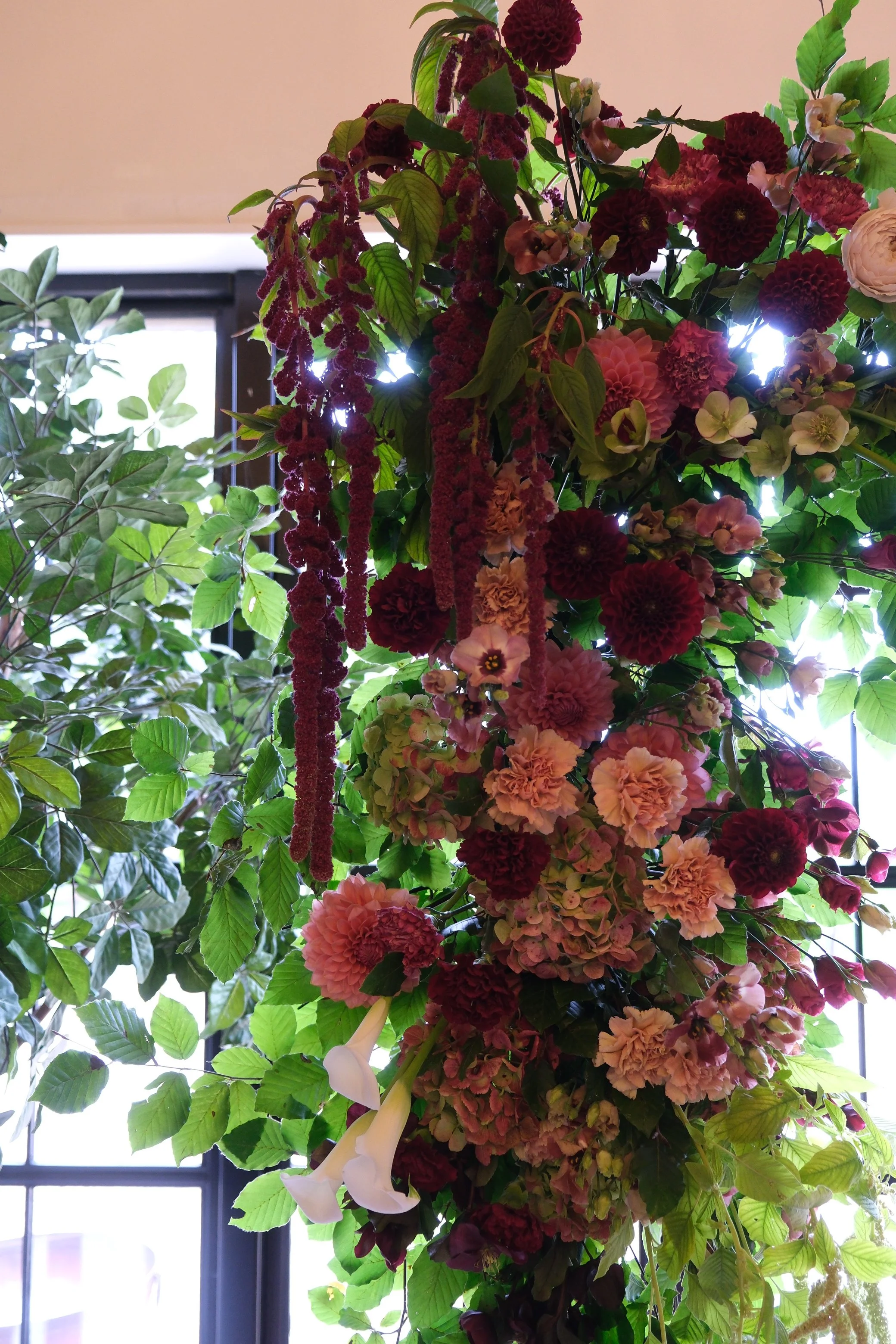 A large floral arrangement with pink, red, and white flowers, green leaves, and hanging dark red flowers, set against a background of windows letting in natural light.