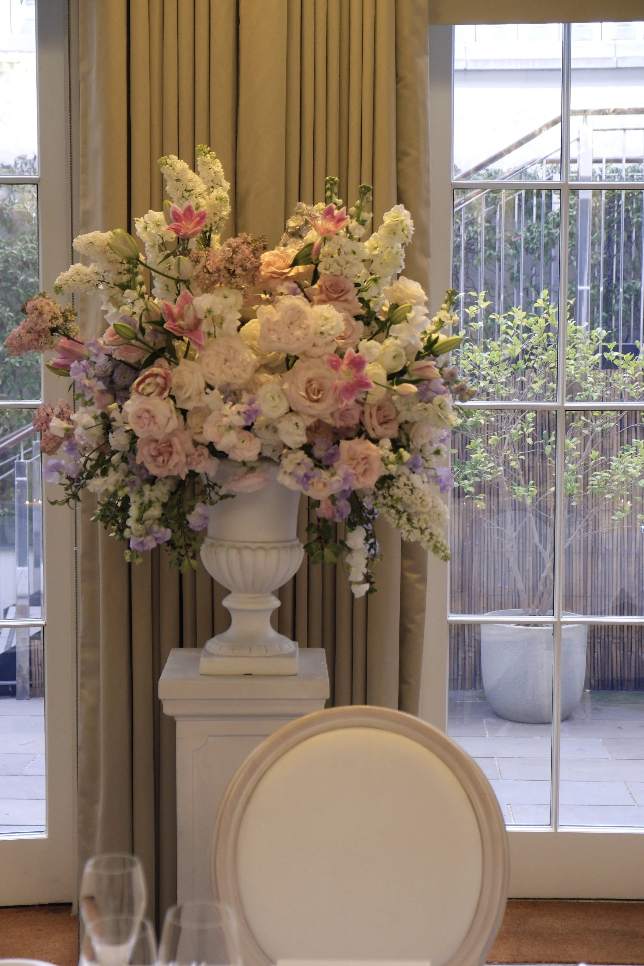 Elegant interior with a large floral arrangement in a white vase on a pedestal, beige curtains, and glass doors showing outdoor greenery and a balcony.