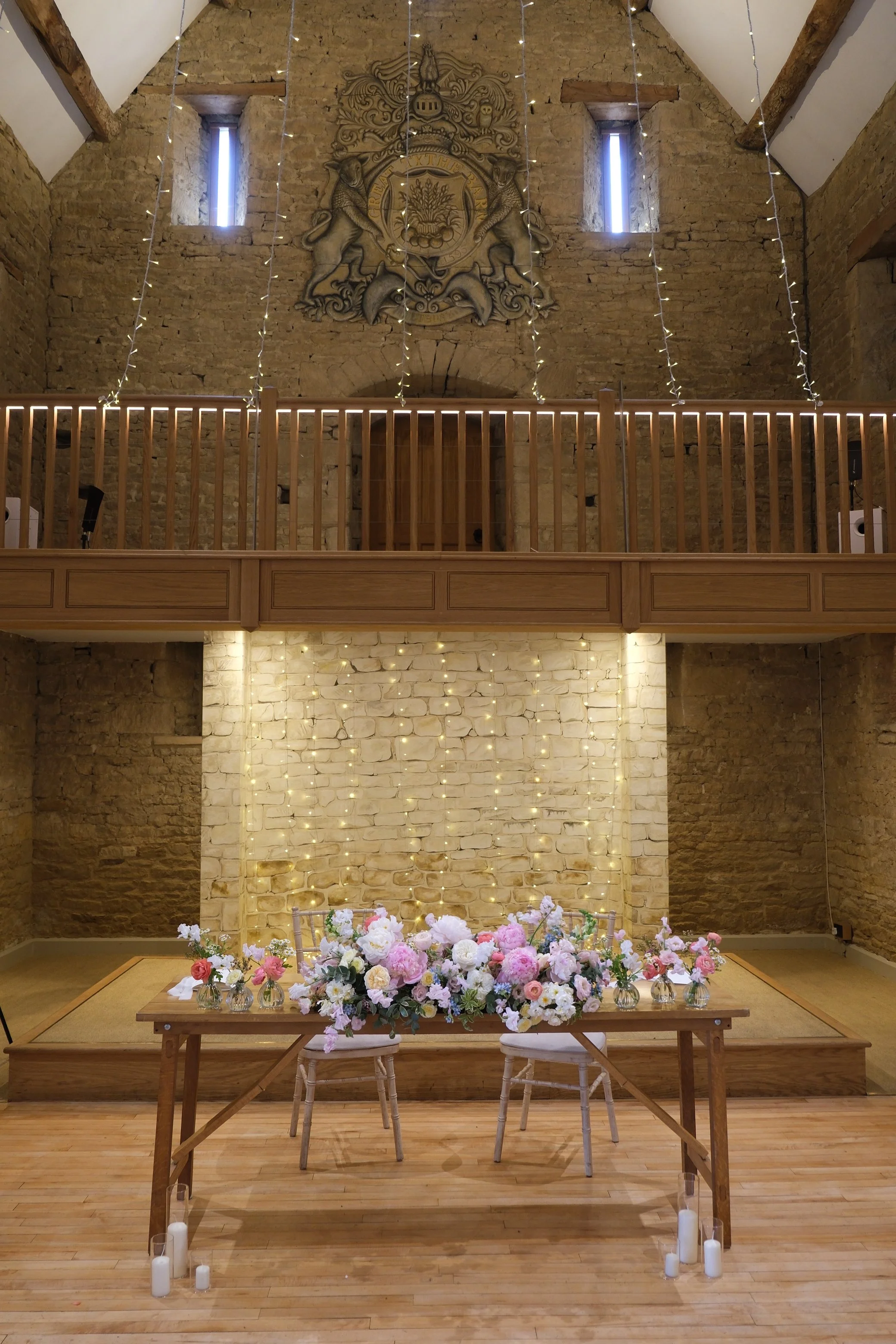Wedding ceremony set up with a wooden table holding a floral arrangement of pink and white flowers, surrounded by glass candle holders with white candles, candles on the wooden floor, a brick wall with string lights and a floral crest, and a wooden balcony railing above.