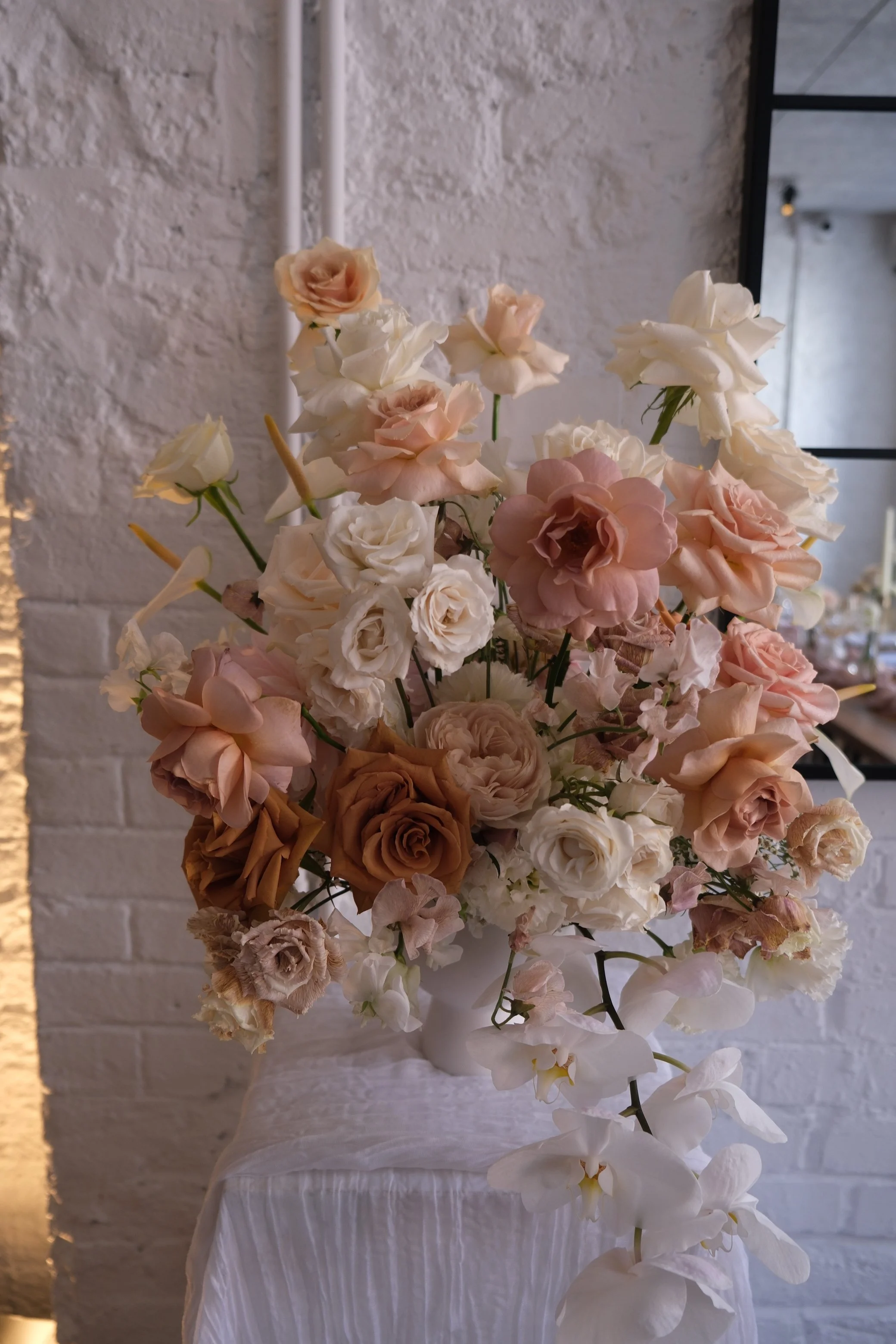 A large bouquet of pastel-colored roses and orchids in a white vase on a white table, with a textured white wall and black-framed mirror in the background.