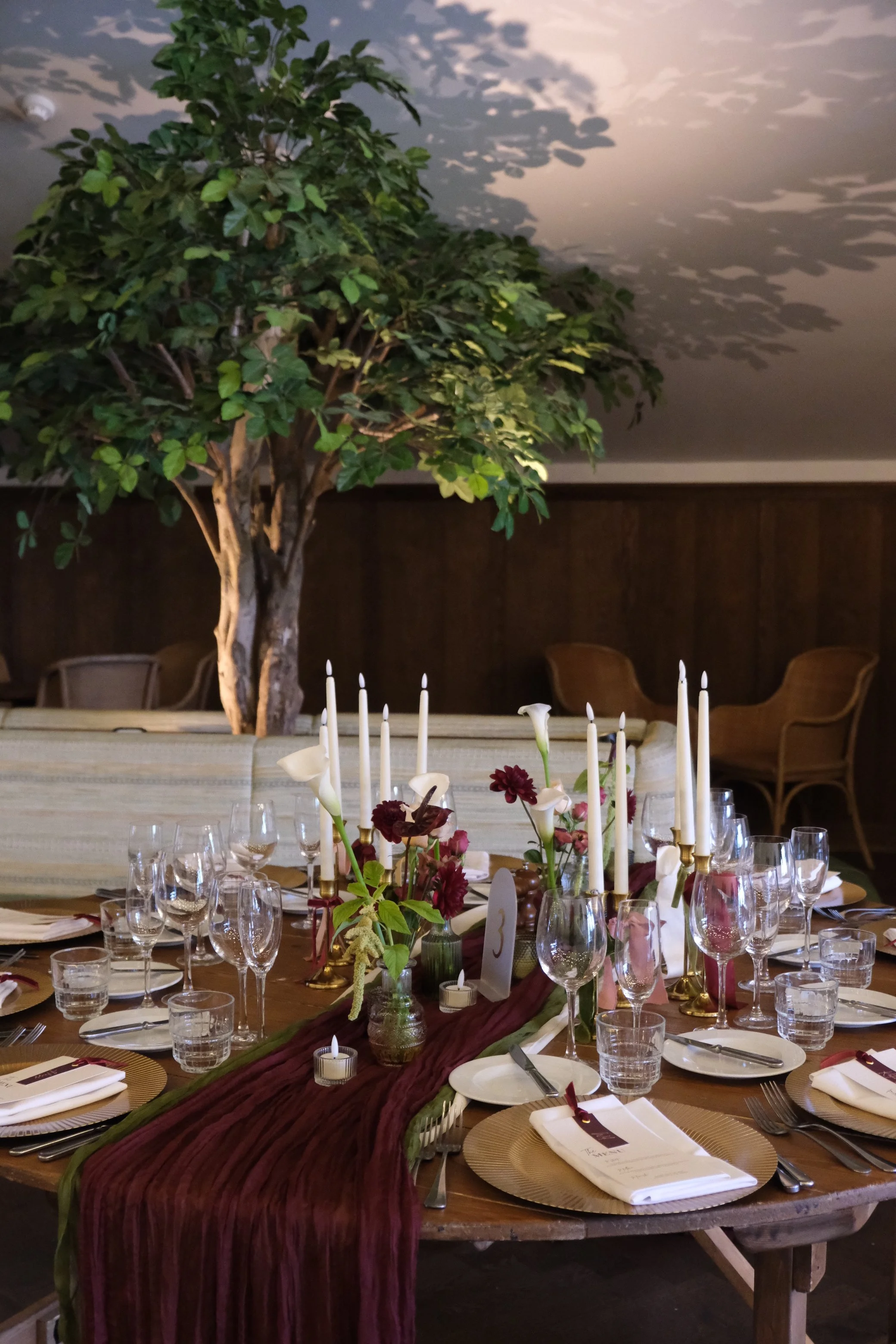 A decorated dining table set for a celebration with flowers and candles, extending beneath a large potted tree in a room with dark wood-paneled walls.