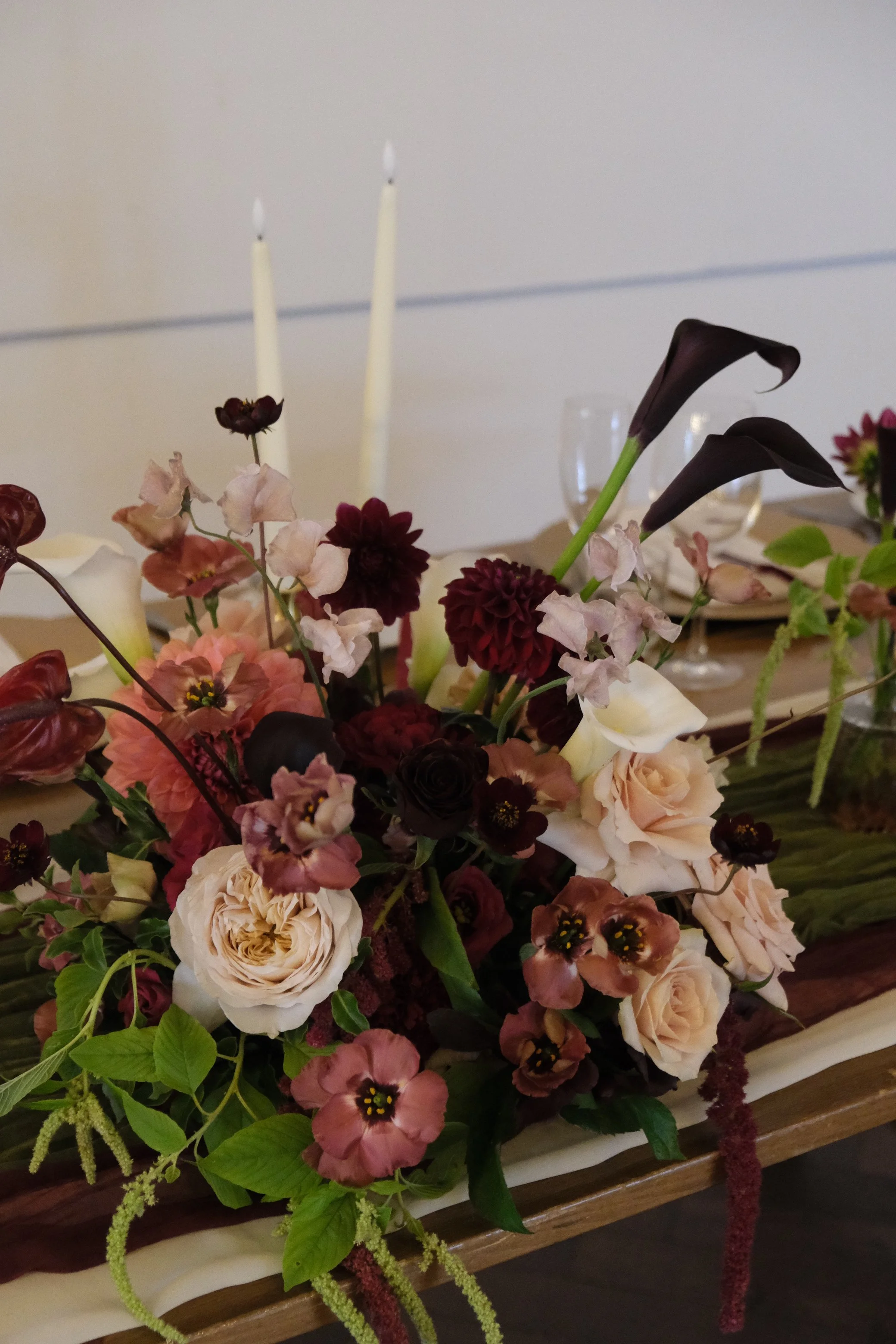 A floral arrangement with dark purple calla lilies, light pink roses, burgundy dahlias, and various greenery, displayed on a table with a white tablecloth and candle holders in the background.