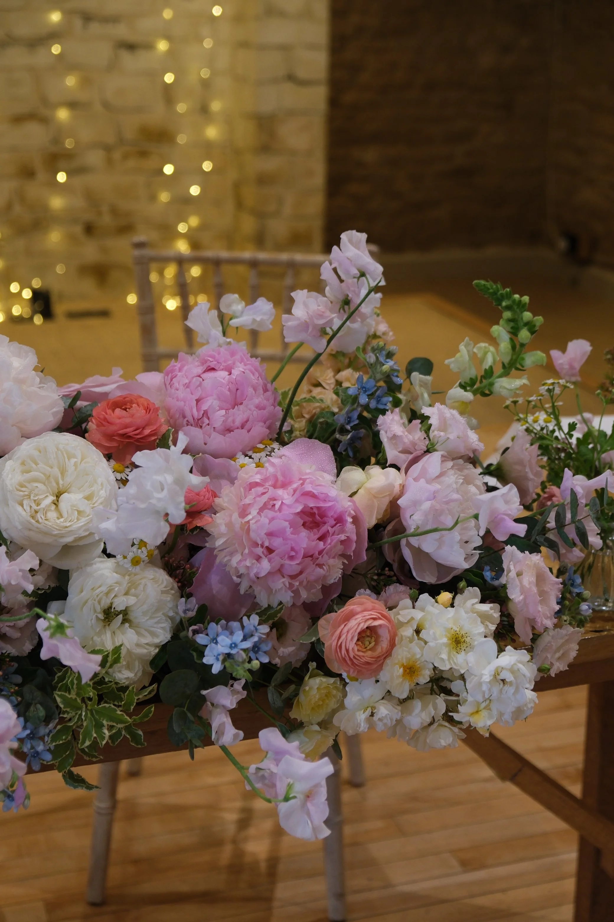 A large bouquet of pink, white, cream, and blue flowers arranged on a wooden table with wooden chairs and a brick wall with string lights in the background.