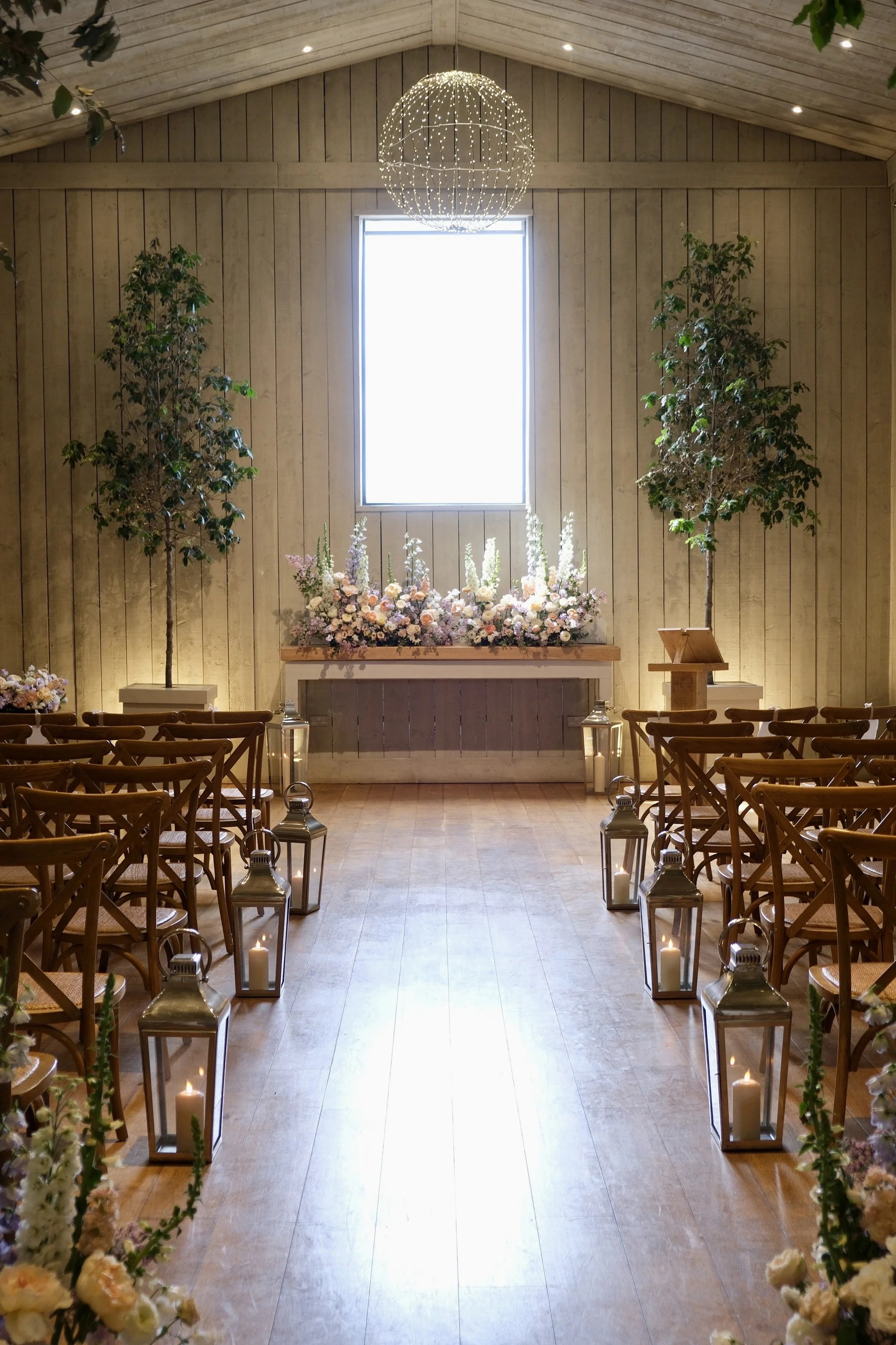 A rustic wedding ceremony space with wooden walls and ceiling. There is a large window at the front, flanked by two potted trees. An LED chandelier hangs from the ceiling, centered above floral arrangements on a table. Wooden chairs are arranged in rows, with glass lanterns and candles lining the aisle.