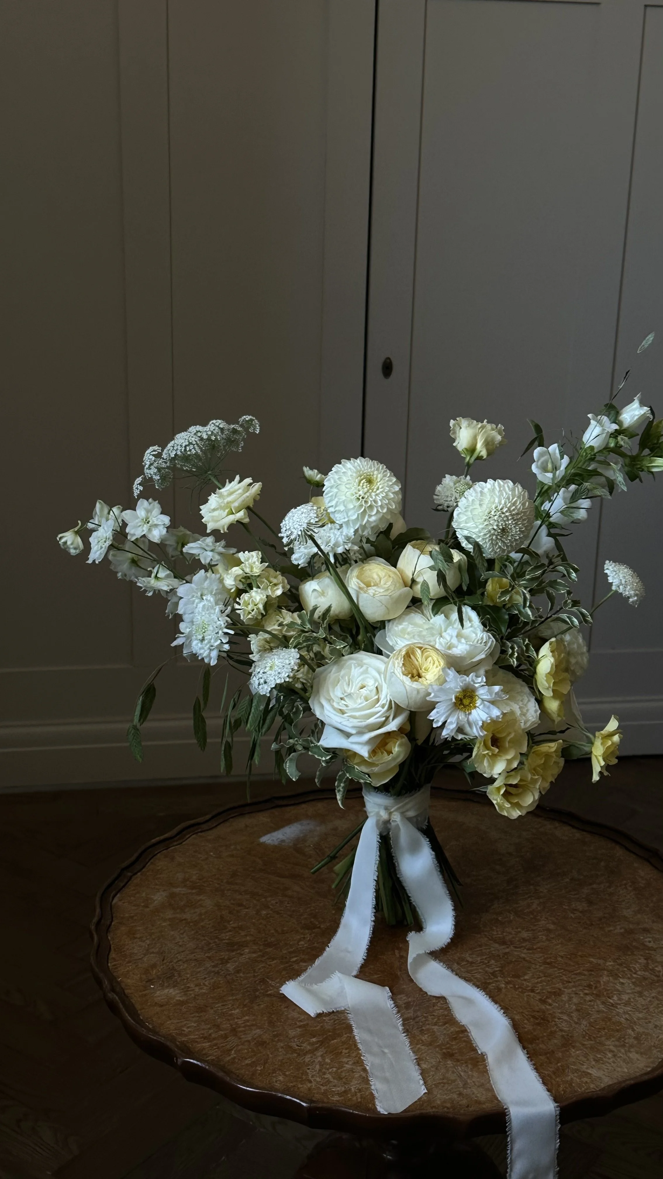 A bouquet of white and cream-colored flowers, including roses, dahlias, and other blooms, tied with a white ribbon, placed on a wooden table in front of a white paneled wall.