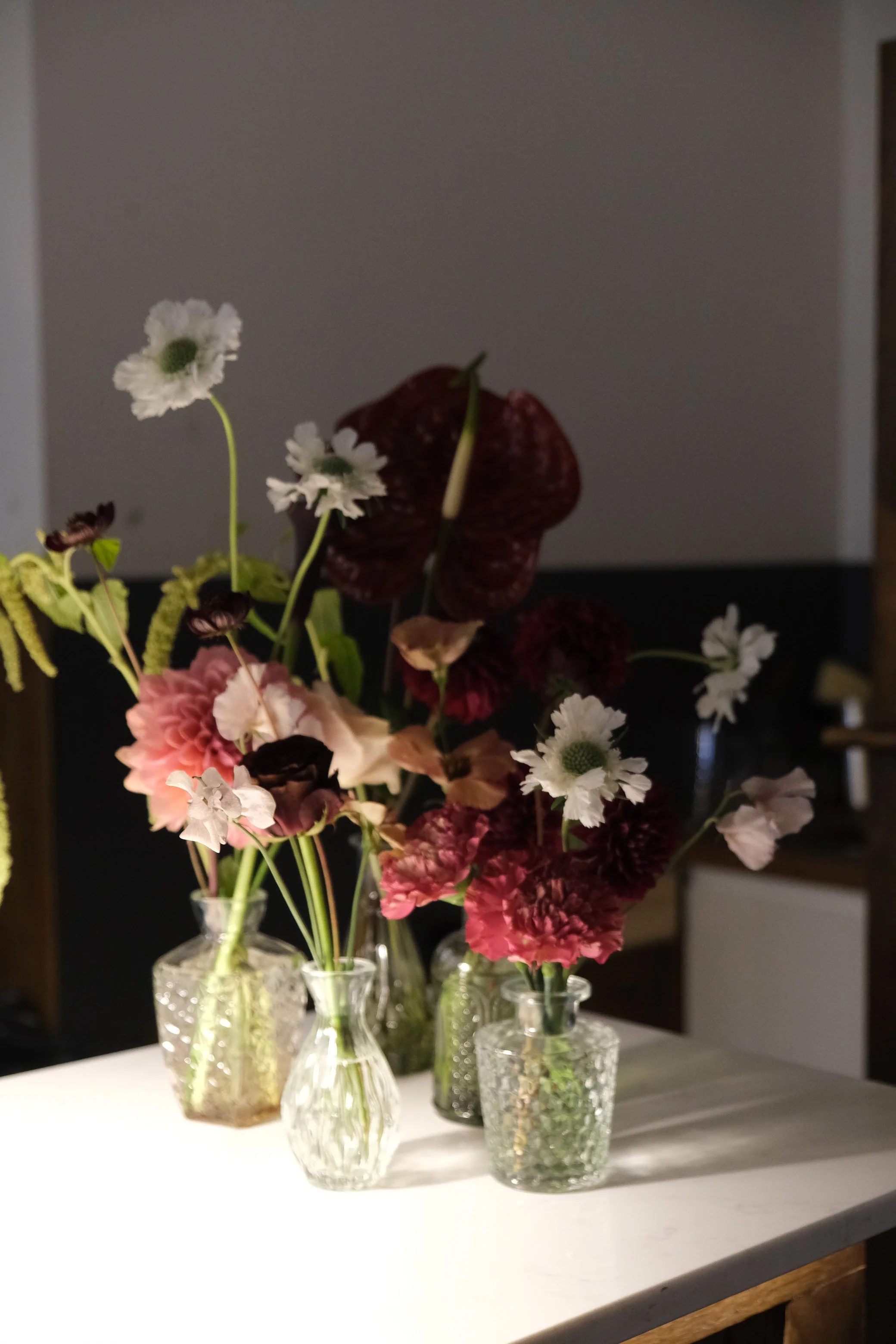 Arrangement of various flowers in multiple clear glass vases on a white table with a dark background.