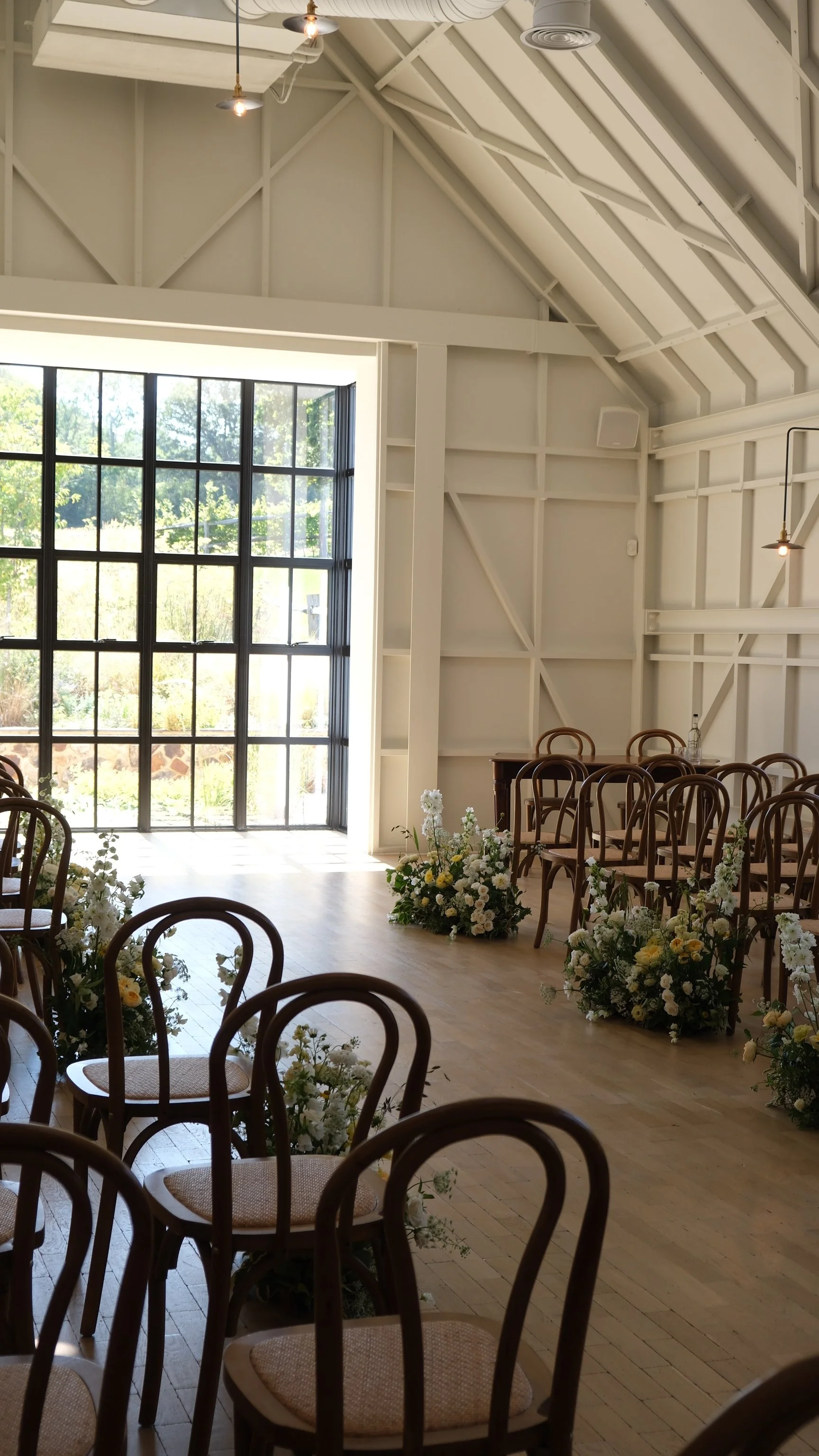 Indoor wedding ceremony setup with wooden chairs arranged in rows, decorated with flowers, and a large window allowing natural light into the room, with a high vaulted ceiling featuring exposed beams.