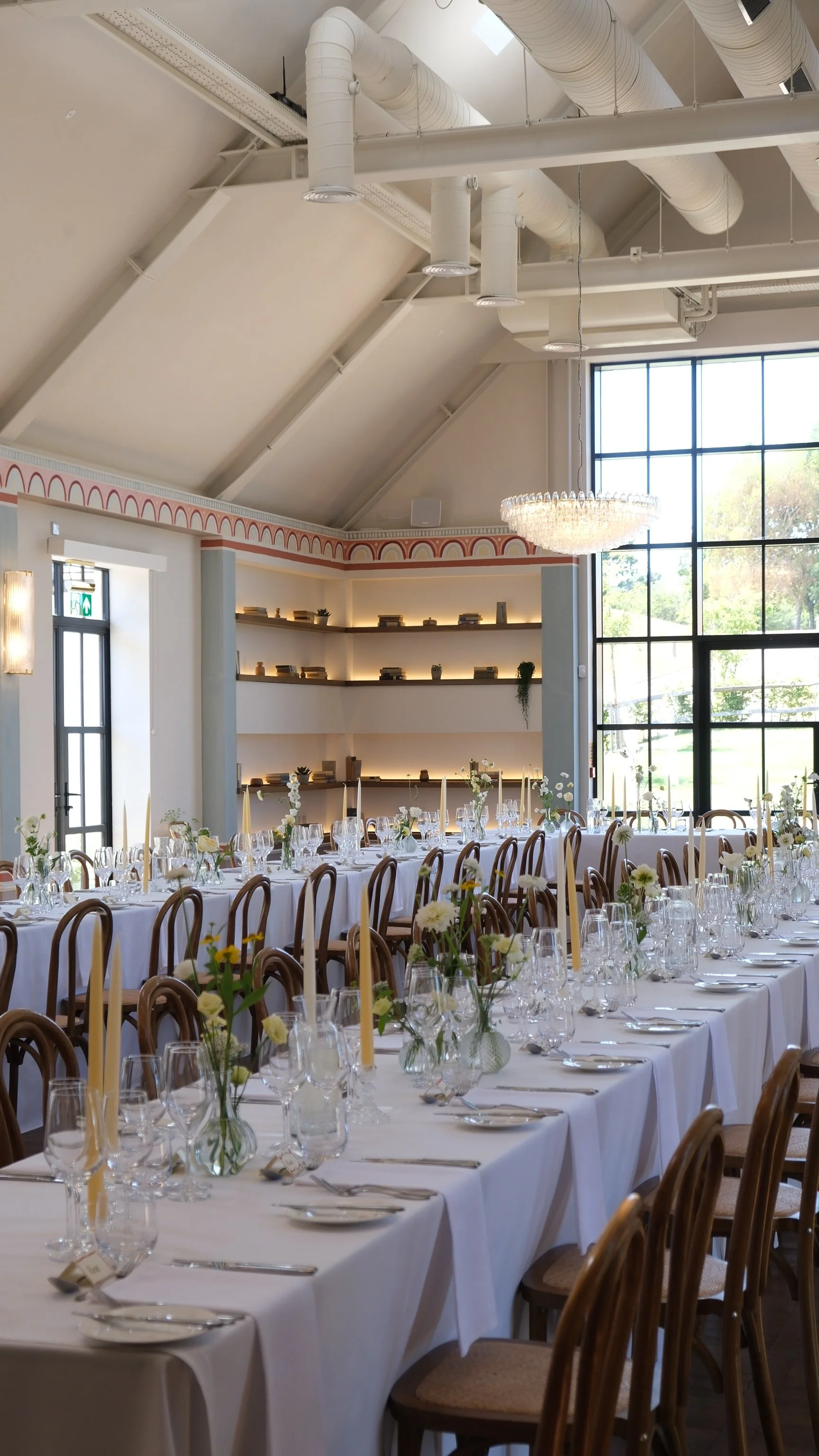 Interior of a decorated banquet hall with long tables covered in white tablecloths, set with glassware, cutlery, and floral centerpieces. The room has large windows, high ceilings with ductwork, a chandelier, and shelves with decor.