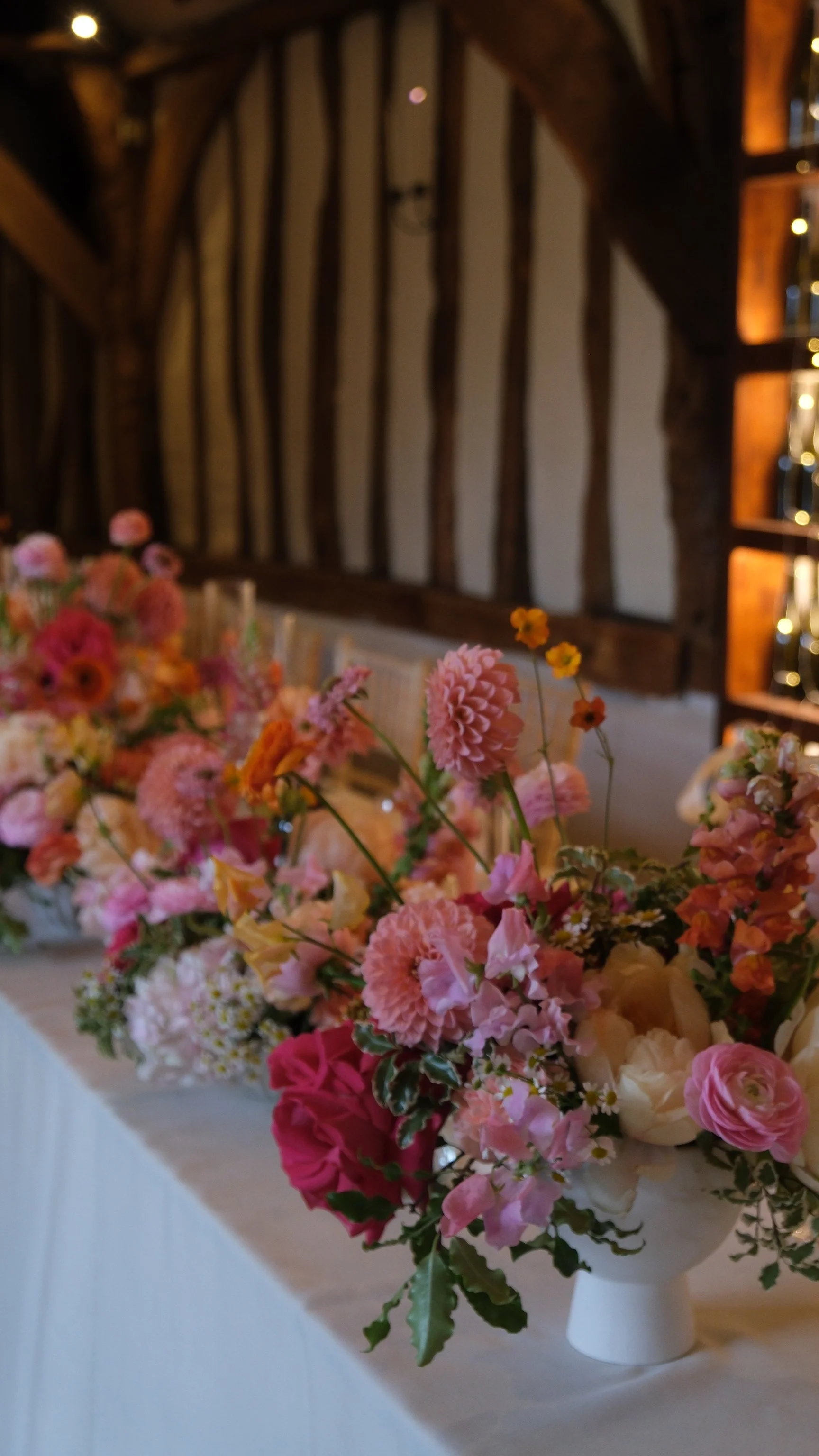 A large floral arrangement with pink, peach, and cream flowers in a white vase, placed on a table with a white tablecloth, in a rustic venue with wood beams and warm lighting.