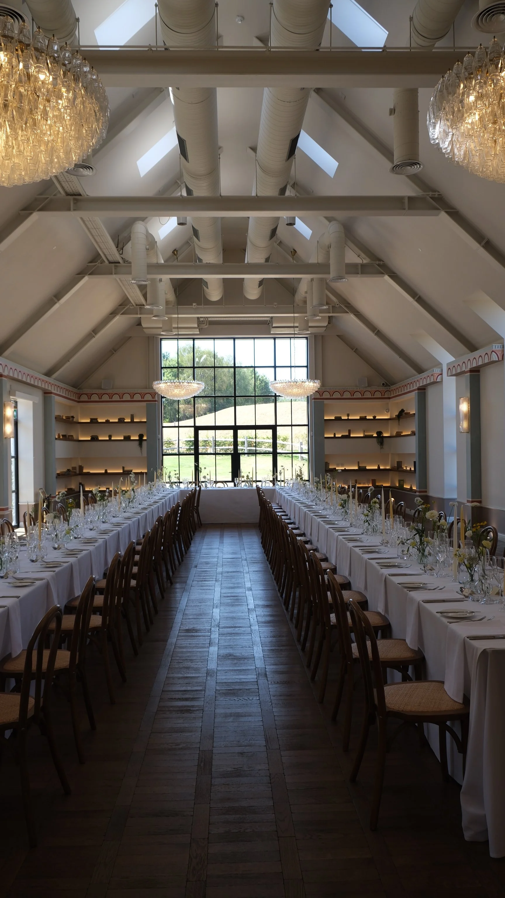 A banquet hall with a long head table covered in a white tablecloth, set with numerous glasses and floral centerpieces, surrounded by wooden chairs. The room features high vaulted ceilings with skylights, large windows at the end, elegant chandeliers, and built-in bookshelves.