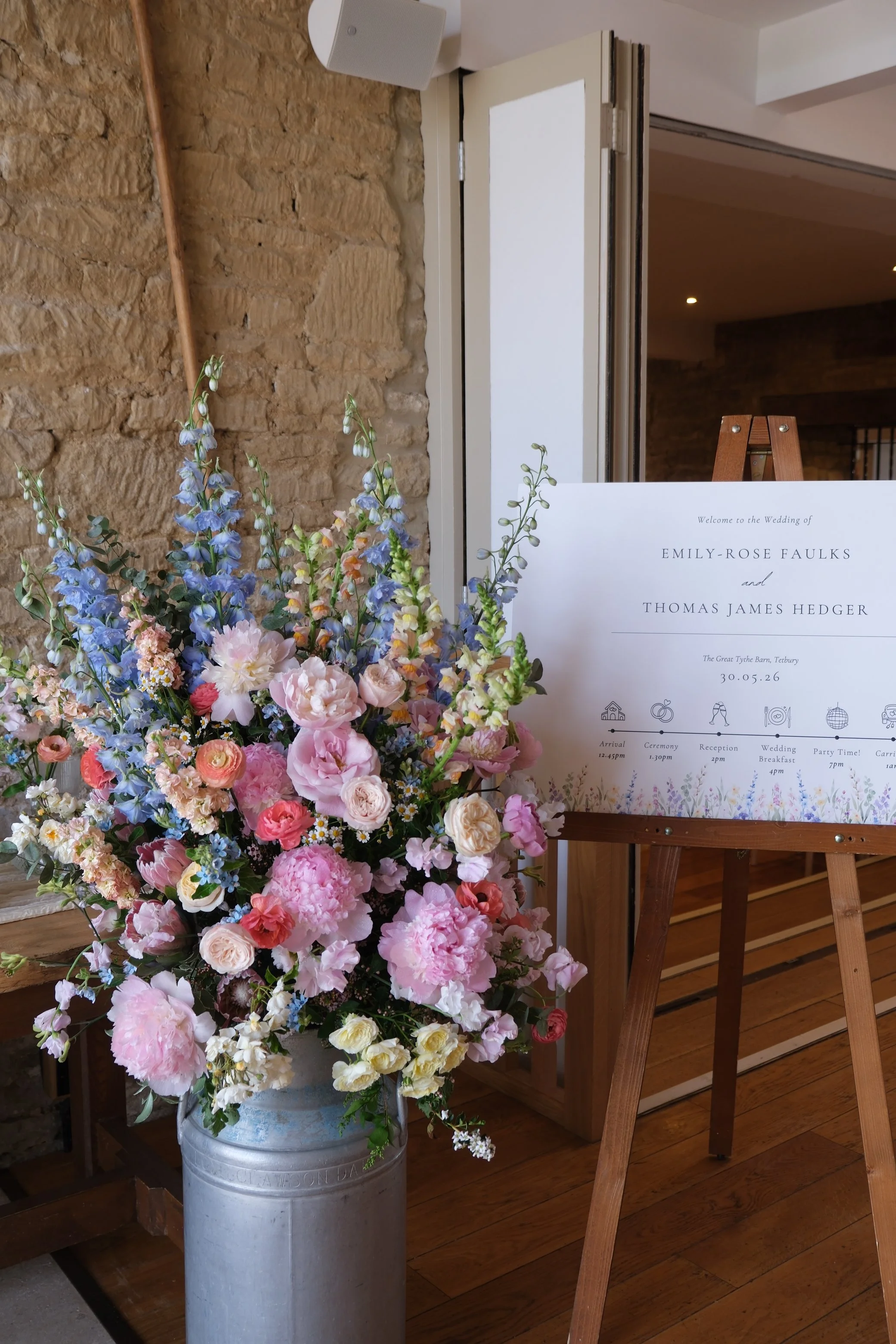 A large arrangement of pink, blue, white, and peach flowers in a grey metal milk churn, with a wedding sign on an easel in the background and a stone wall to the side.
