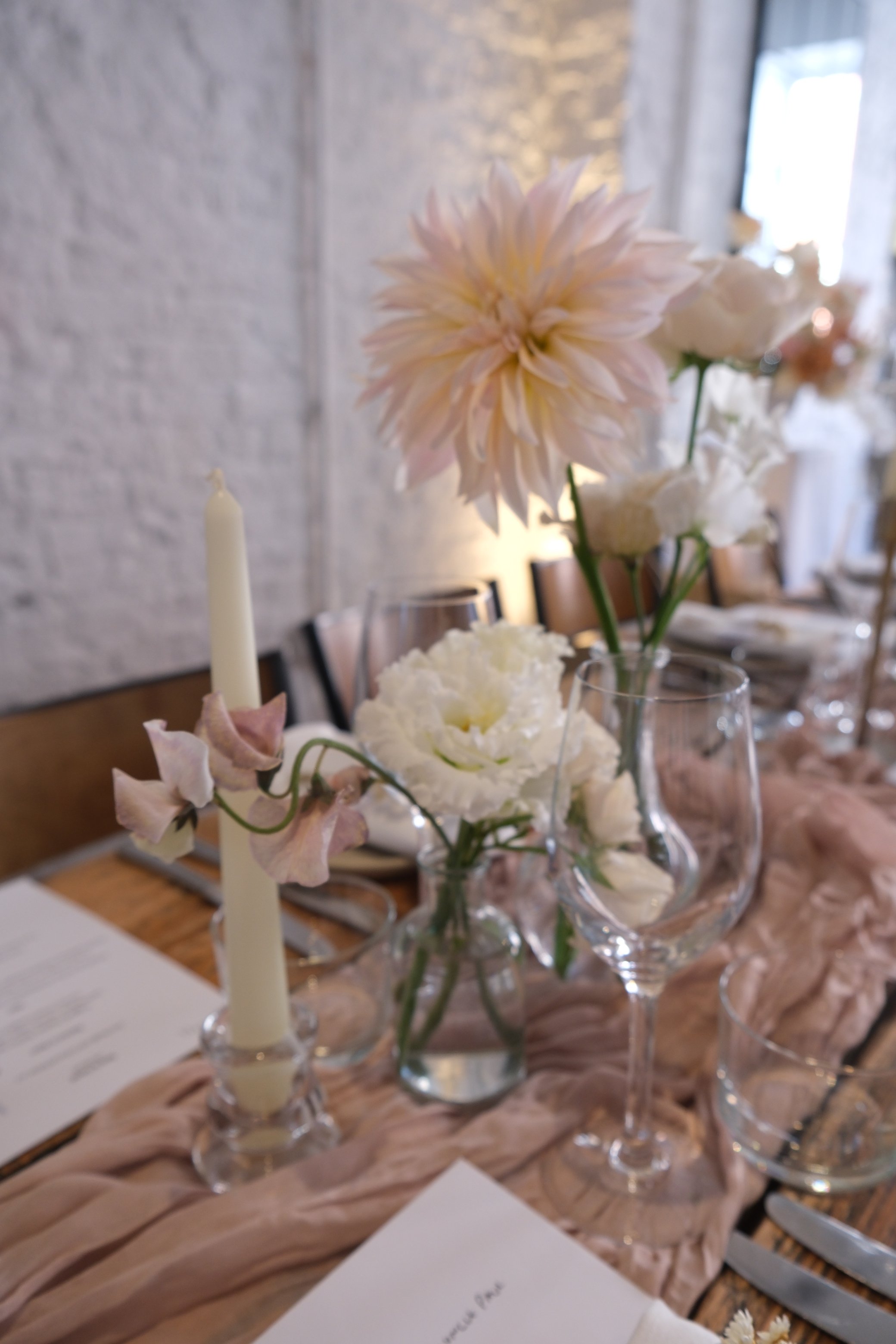 A table decorated with various flowers in glass vases, including a large light pink dahlia, surrounded by white flowers and a candle, set against a white brick wall and window in the background.