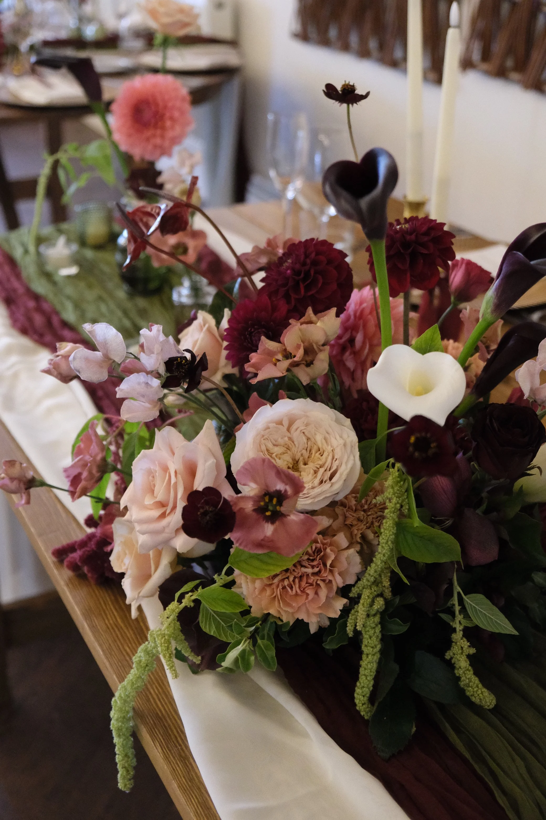 A floral arrangement with pink, burgundy, white, and dark purple flowers, accented with green foliage, set on a wooden surface with a white cloth draped underneath. In the background, there are glasses and a blurred dining setting.
