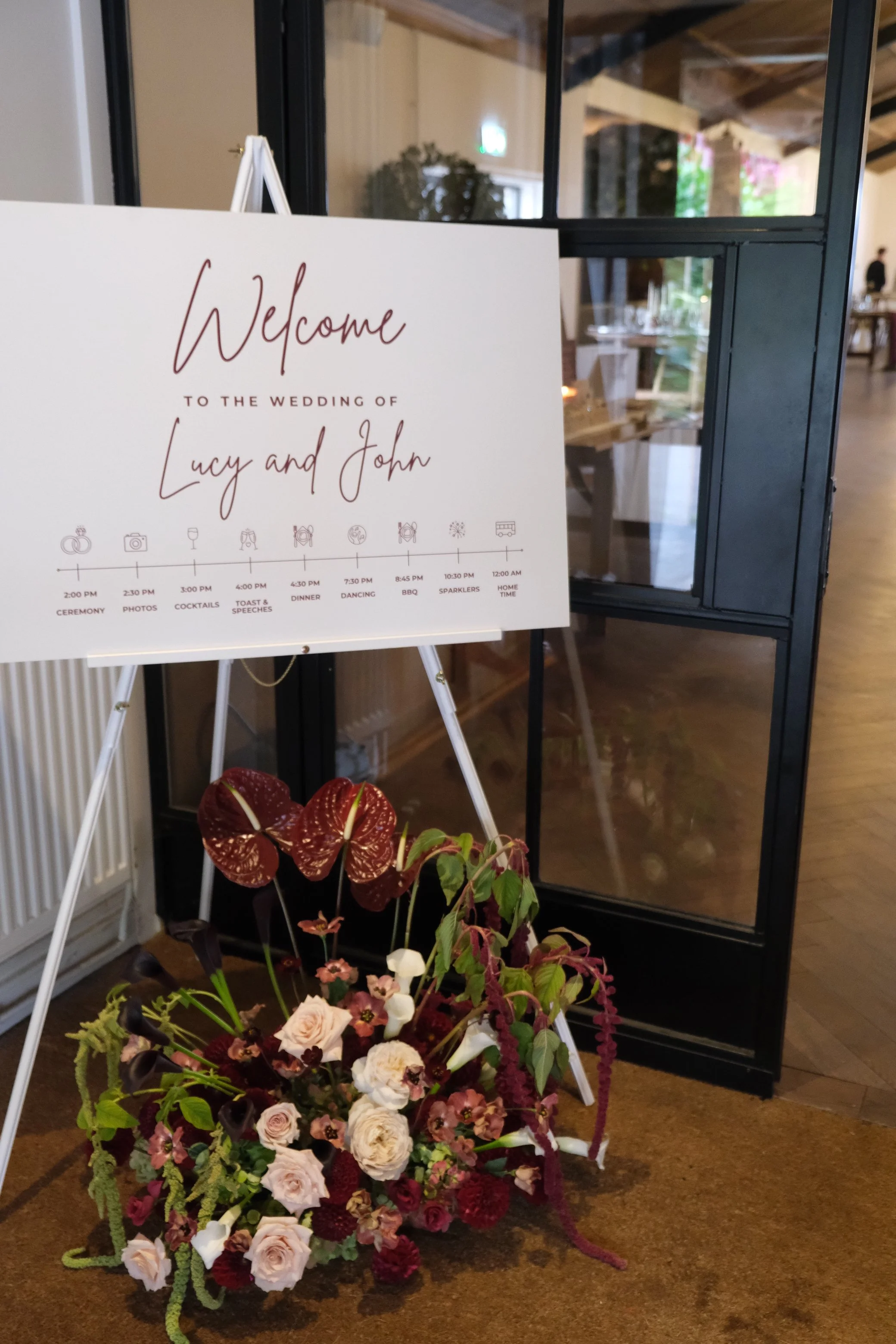 Wedding reception sign with schedule and floral arrangement in front of a glass display case inside a rustic venue.