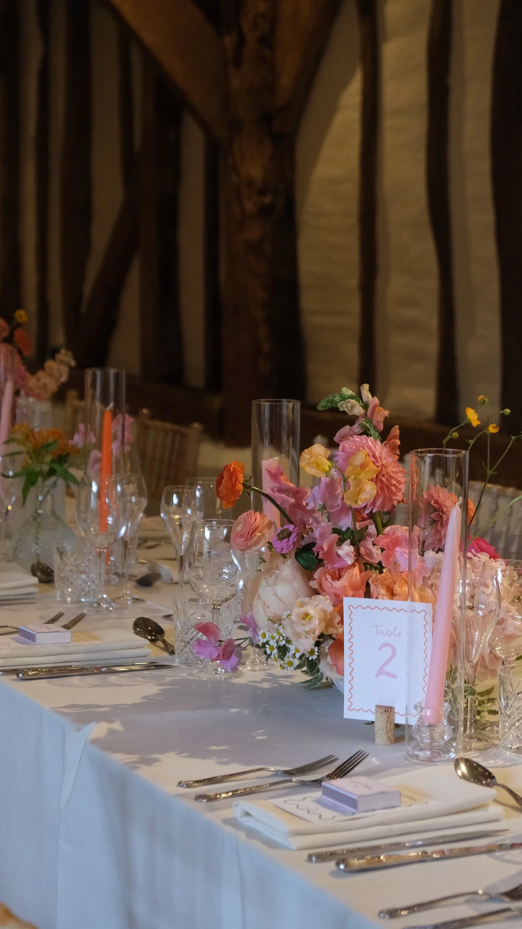 A banquet table decorated with a pink floral centerpiece, surrounded by candles, glassware, and tableware, with a rustic wooden wall in the background.