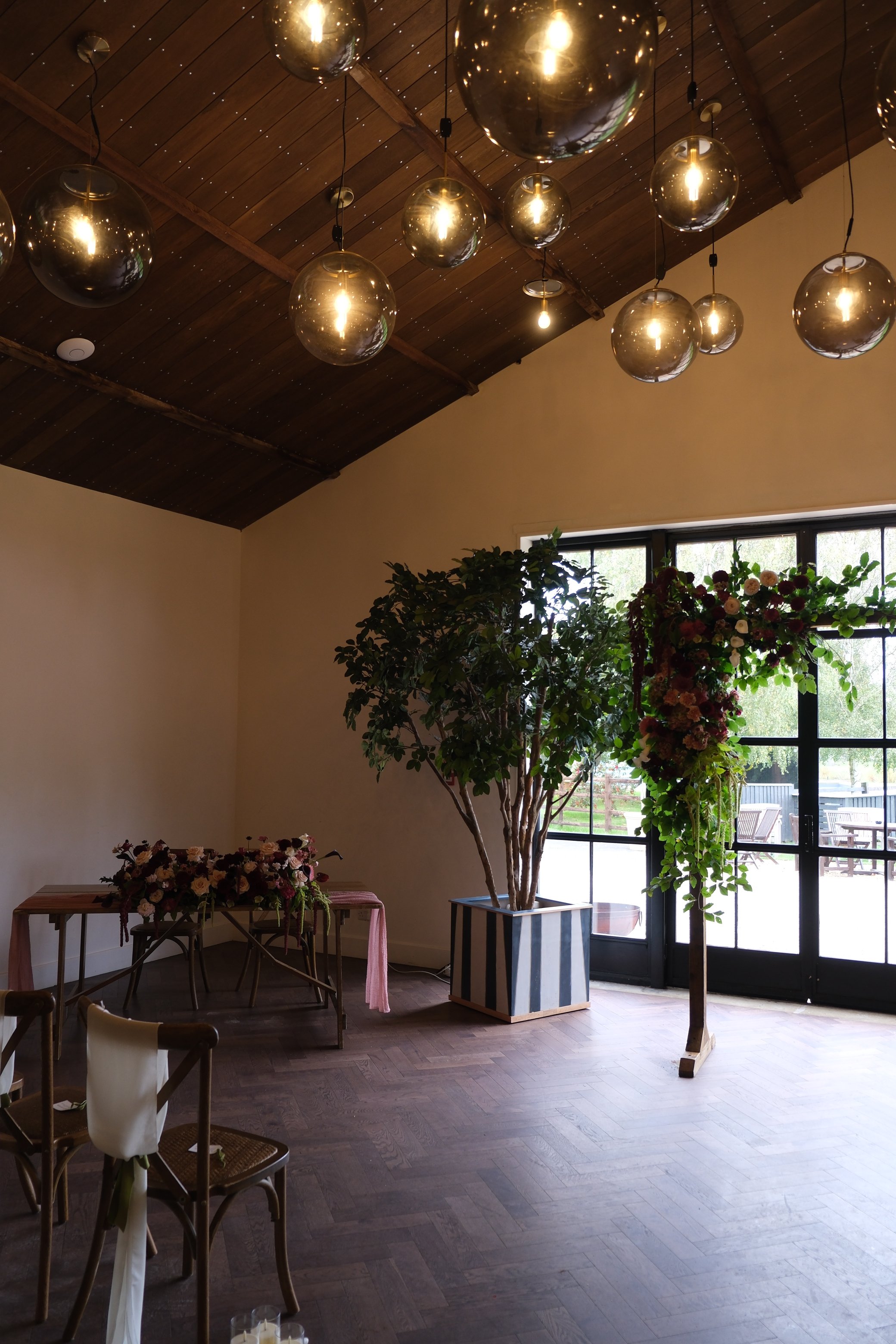 Interior of a room with a wooden ceiling featuring multiple hanging spherical pendant lights, large windows with black frames, a potted tree in a striped planter, and tables decorated with floral arrangements.