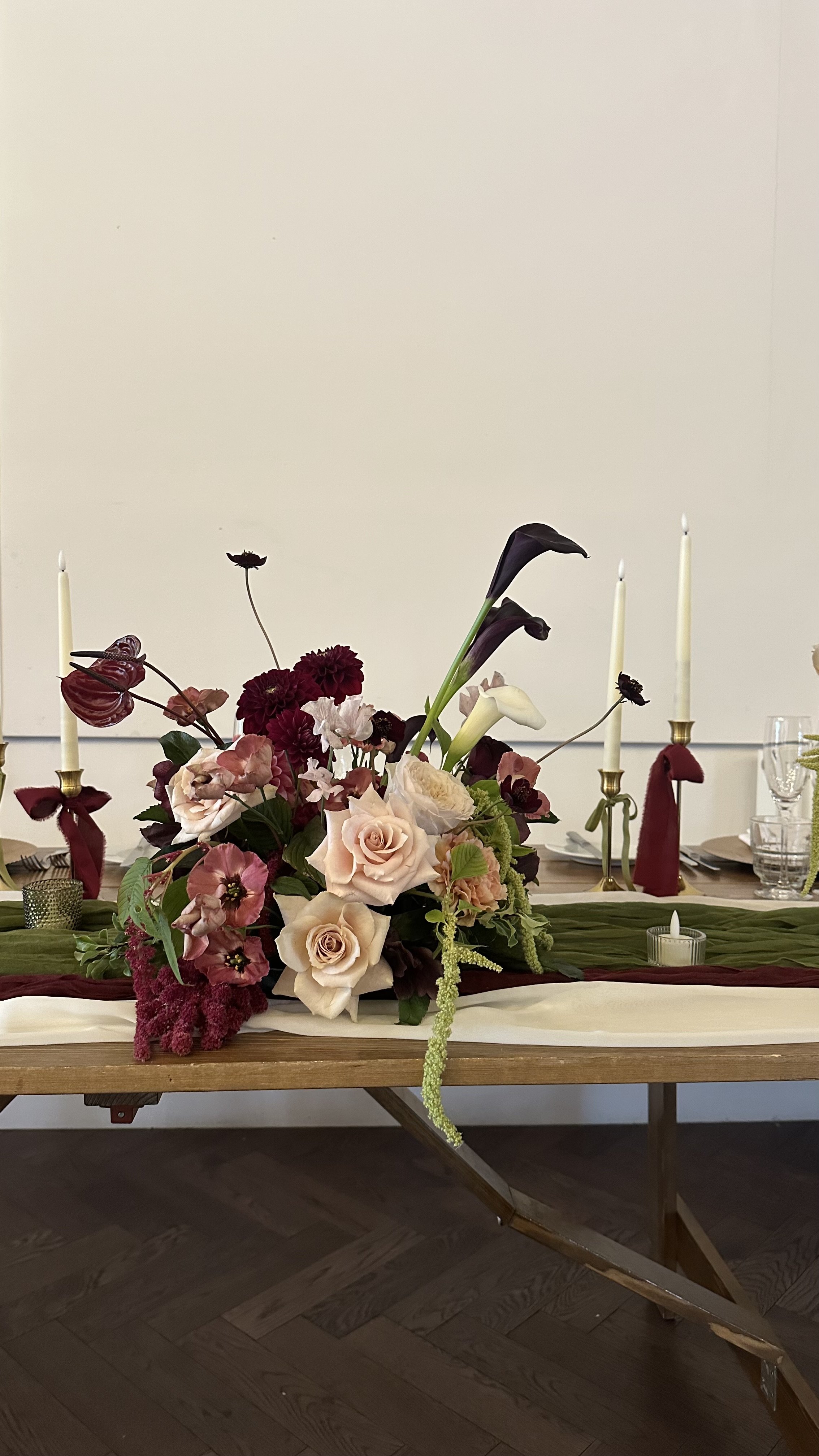 Elegant table centerpiece with various flowers including roses, calla lilies, and dark red blooms, flanked by tall white candles with red and burgundy ribbons, on a wooden table.