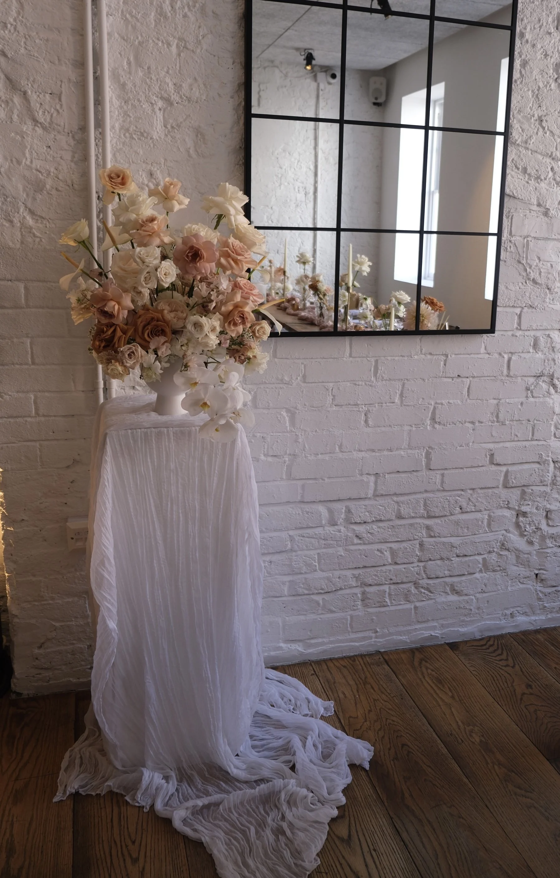 A white flower arrangement in a white vase placed on a table covered with a white cloth, in front of a white brick wall with a large mirror reflecting a window and more flowers.