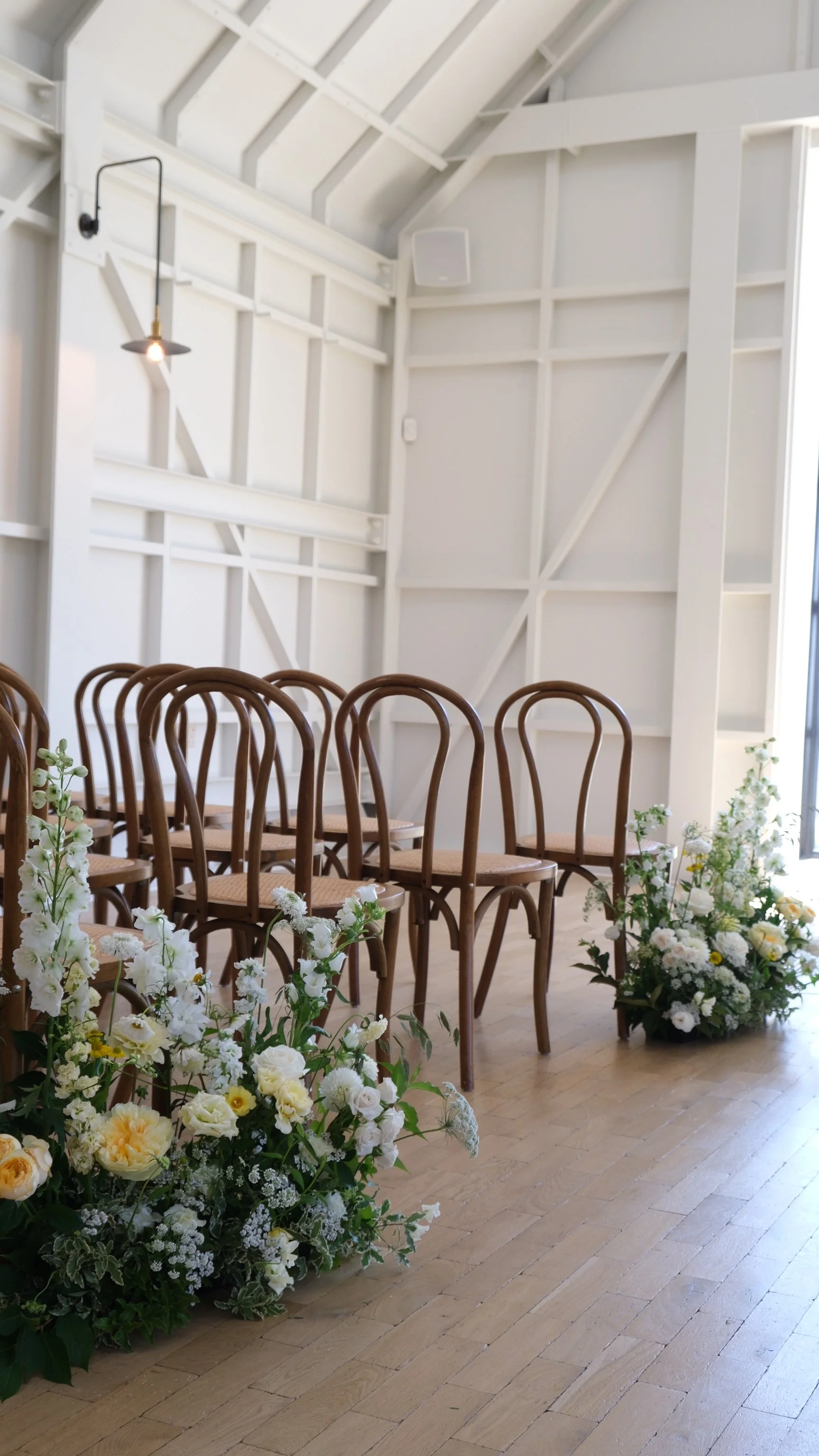 Interior space with flower arrangements and wooden chairs, likely set up for a wedding or event, against white paneled walls with a single hanging light fixture.