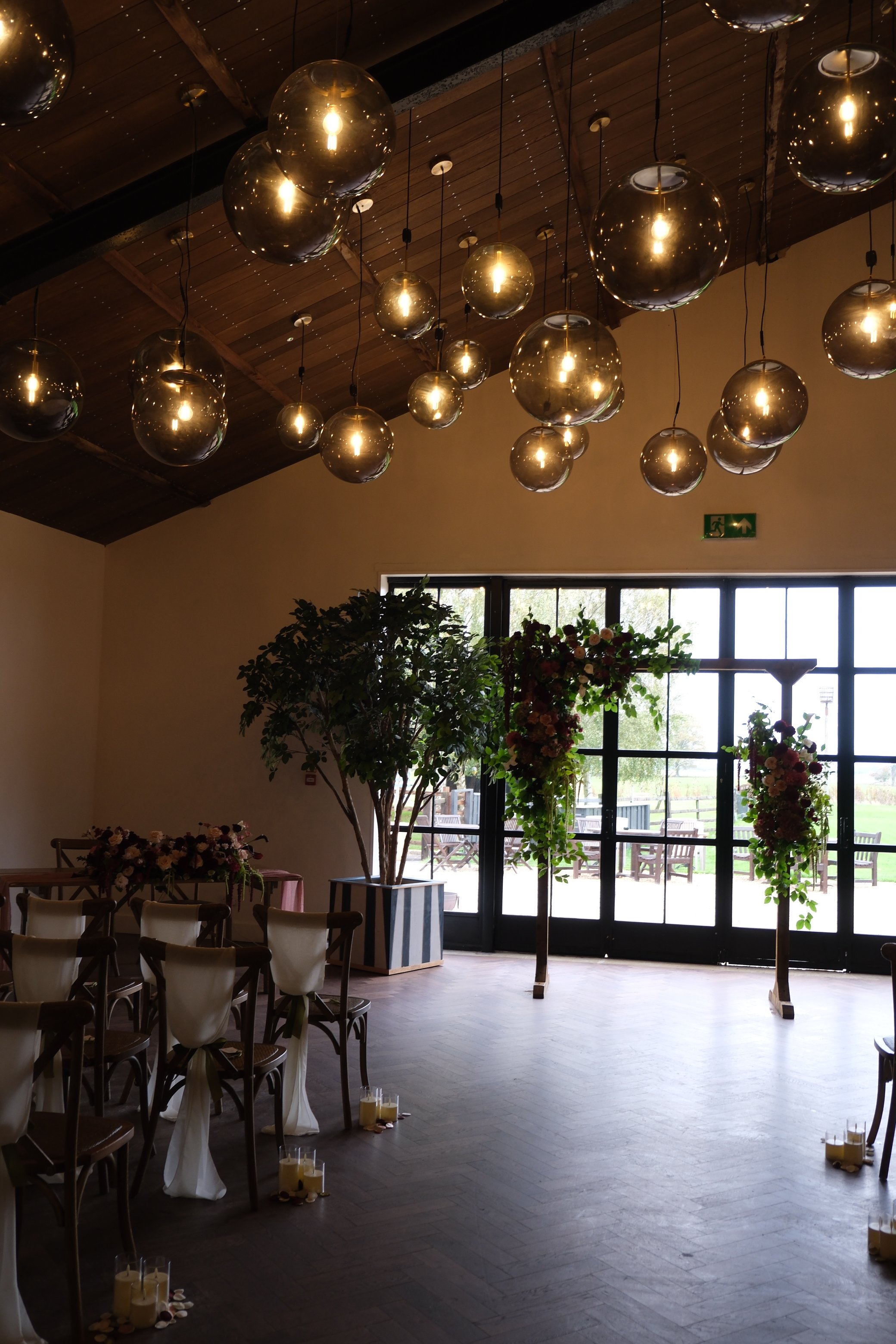 Interior of a banquet hall decorated for a wedding with hanging glass sphere pendant lights, floral arrangements, candles on the floor, and large windows with outdoor seating visible.