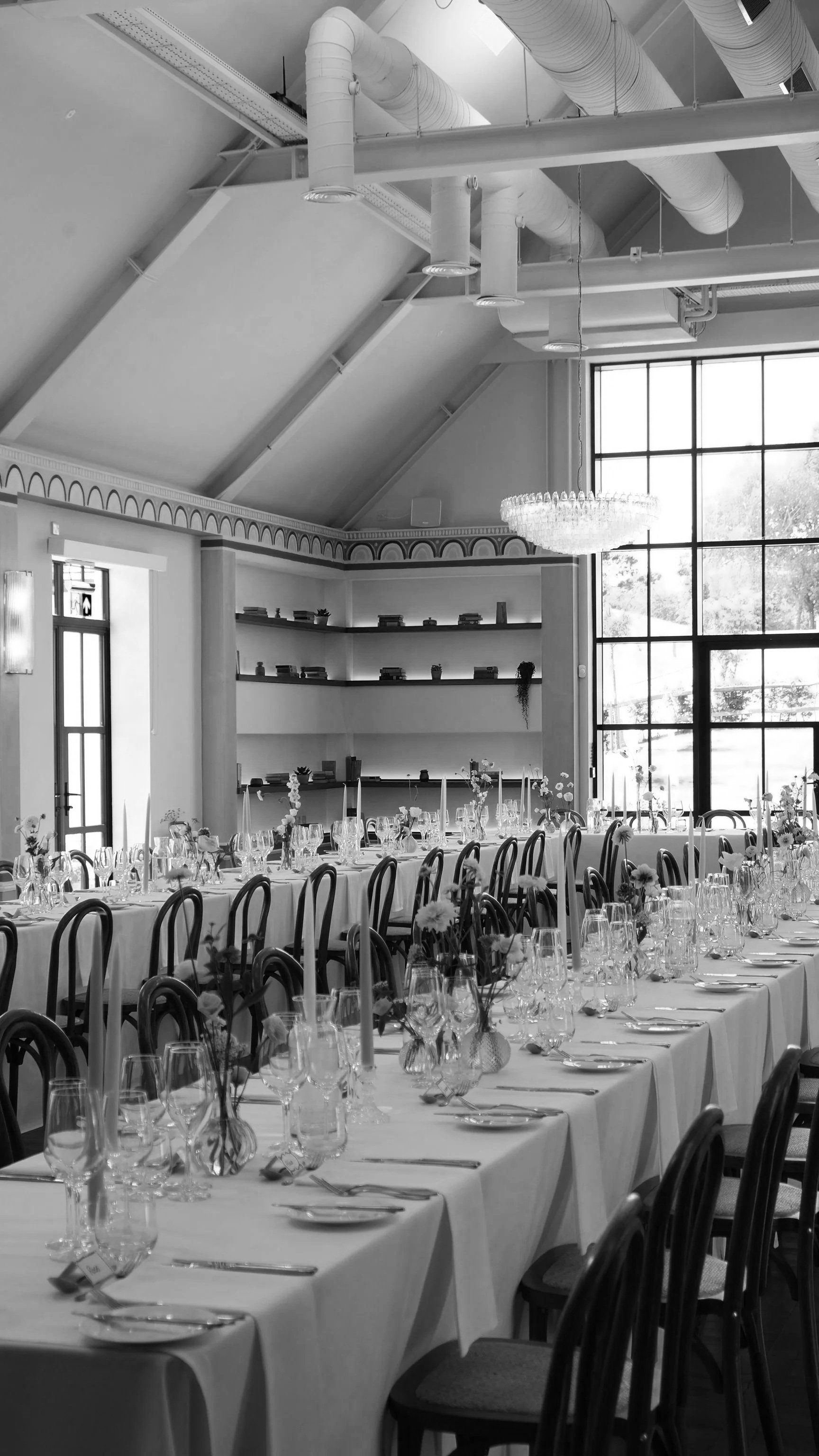 Elegant banquet room with long tables set for a formal event, decorated with flowers and glassware, large windows letting in natural light, and a chandelier hanging from the high ceiling.